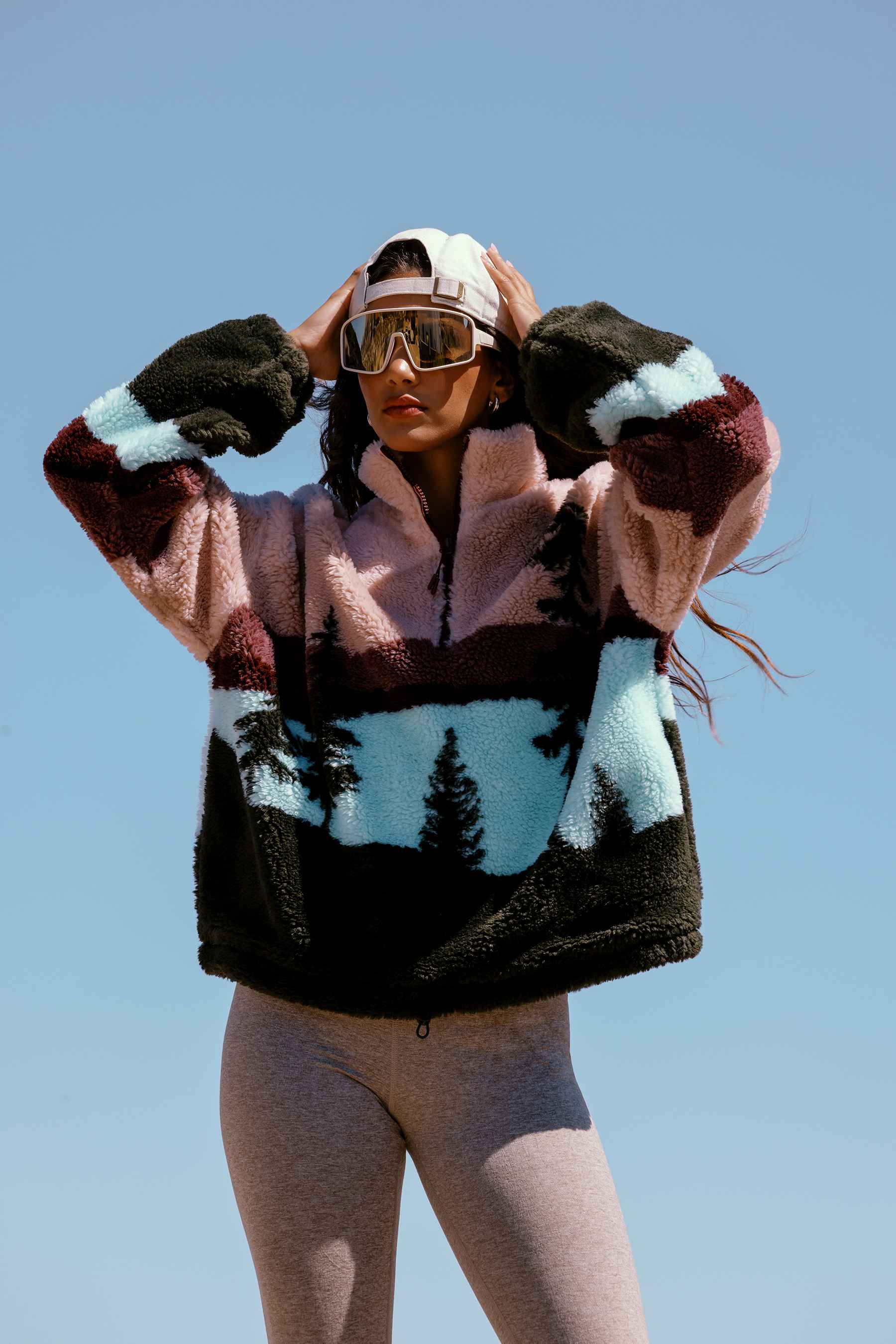 A woman wearing a fuzzy color-blocked fleece pullover with a scenic pine tree pattern, ski goggles, and a white cap against a clear blue sky.