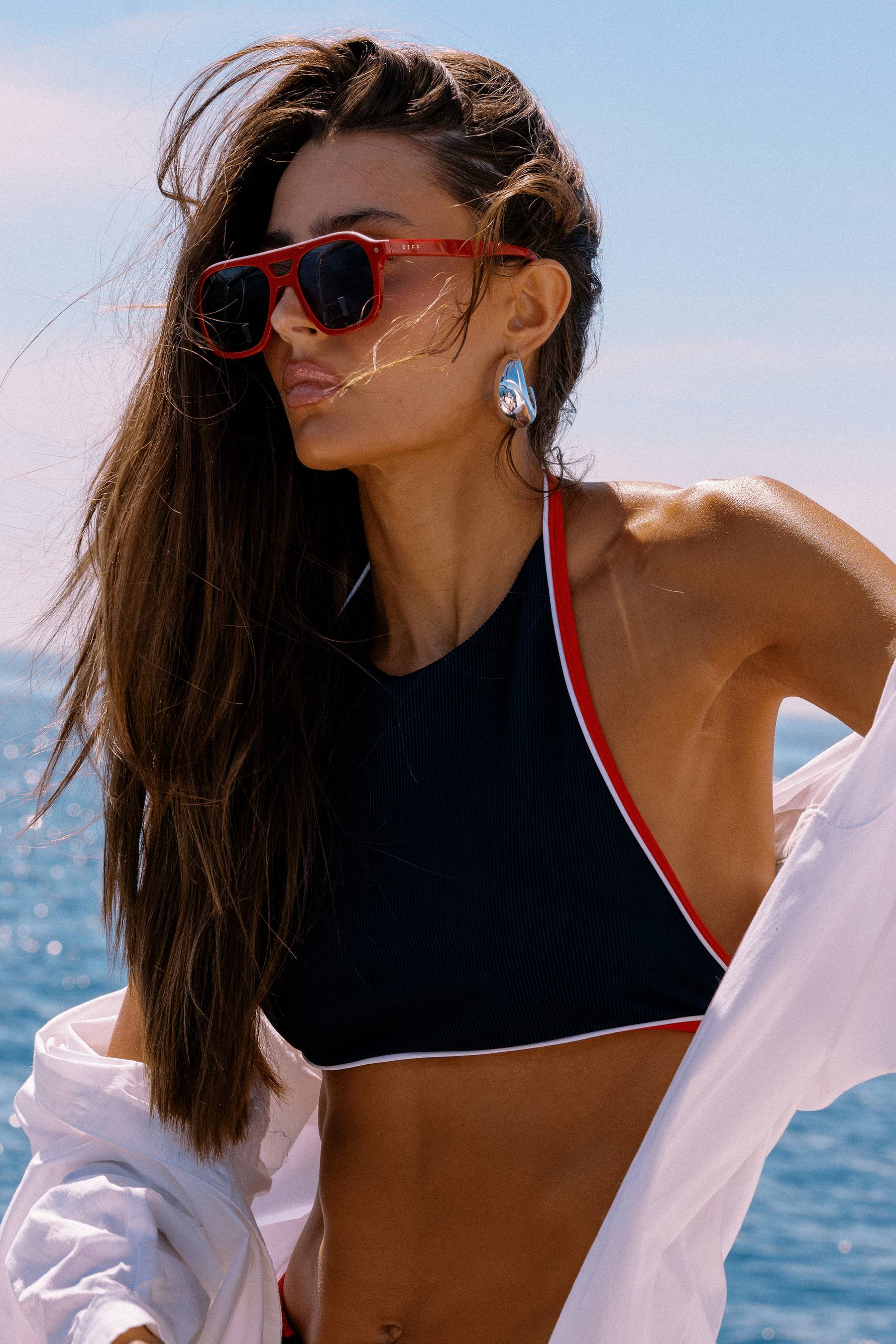 Woman wearing a black bikini with red straps and sunglasses, standing by the water.