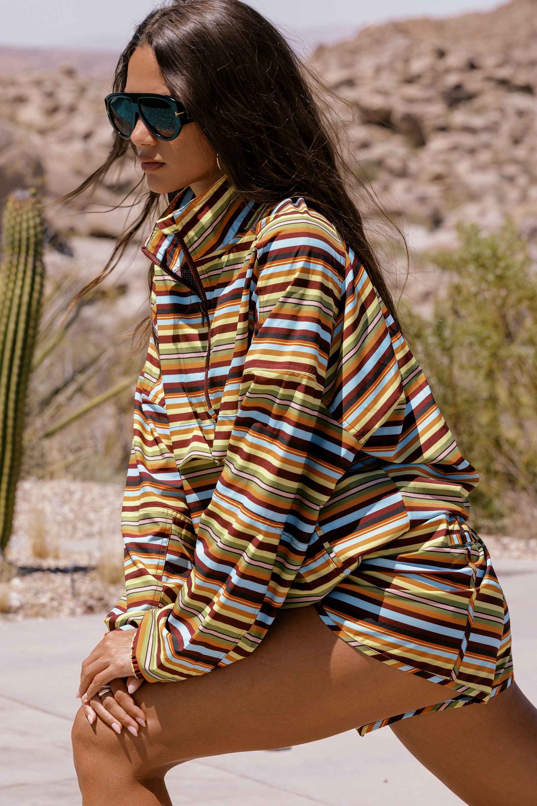 A woman in a desert setting wearing a colorful striped long-sleeve shirt and matching shorts, paired with dark sunglasses.