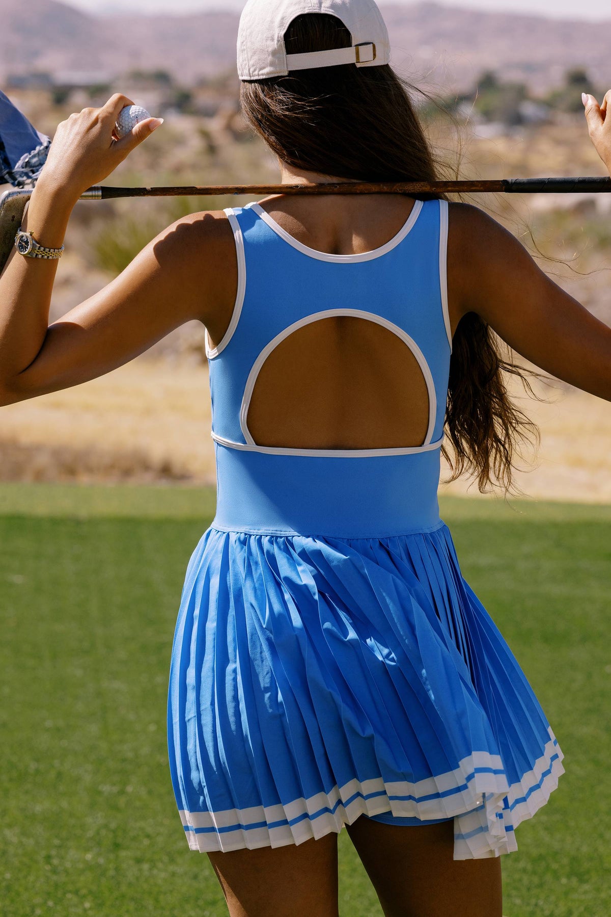 A woman on a golf course seen from behind wearing a light blue sleeveless athletic top with a large open back and a matching pleated blue skirt with white trim, holding a golf club over her shoulder.