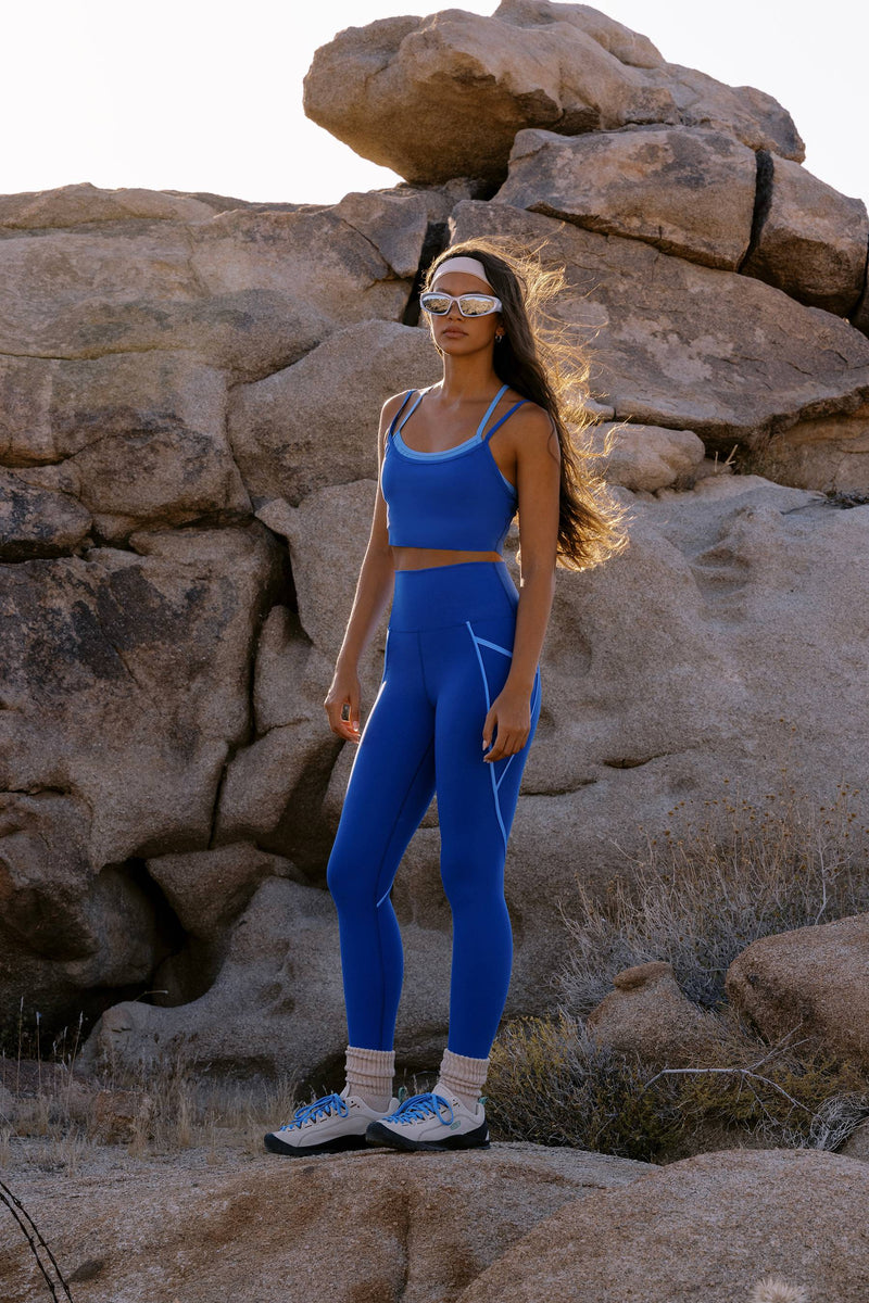 A woman wearing a matching blue athletic crop top and leggings set with white trim stands outdoors in front of large rock formations, wearing white sneakers and white-framed sunglasses.
