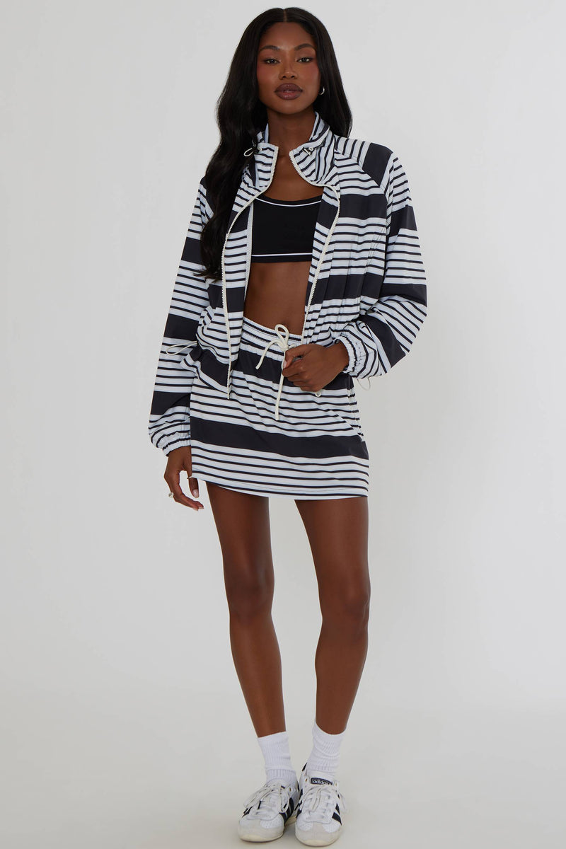 A model stands against a light gray studio background wearing a matching black and white horizontally striped zip-up jacket and mini skirt set, styled with a black crop top, white socks, and white sneakers.