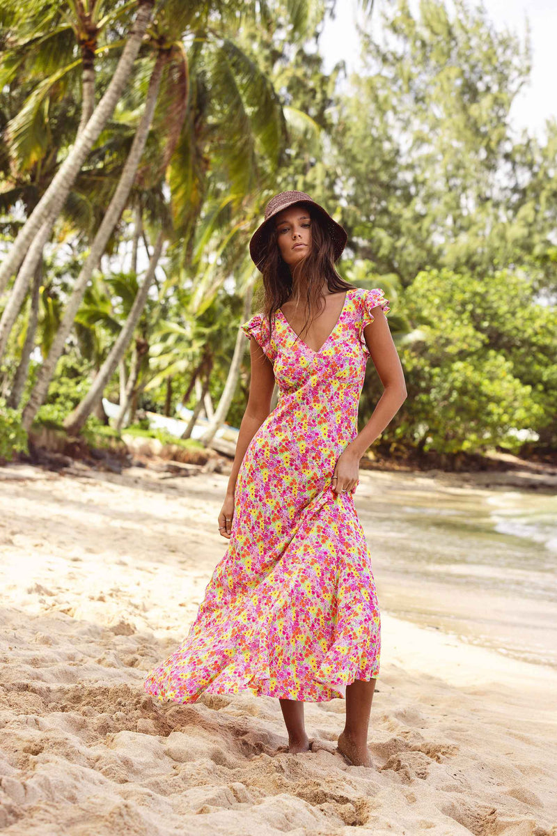 Woman in a floral dress and hat standing on a sandy beach with palm trees in the background
