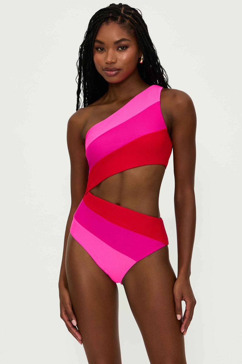 A model wearing a one-shoulder asymmetrical one-piece swimsuit in red and pink color blocks with a cutout detail at the waist.