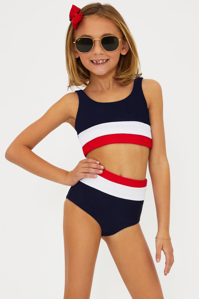 A girl wearing a two-piece swimsuit with an Americana colorblock pattern, consisting of red, white, and blue colors. She is also wearing sunglasses and there is a bow in her hair.