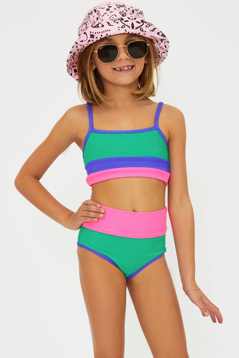A young girl wearing a color-blocked swimsuit with green, pink, and purple colors. She is also wearing a patterned hat and sunglasses.