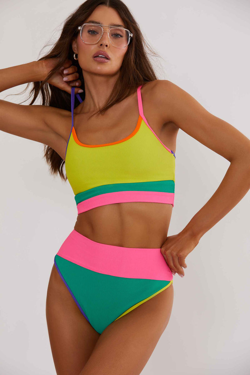 A woman wearing a multicolor block swimsuit with yellow, pink, and green colors. The swimsuit has adjustable straps with gold hardware.