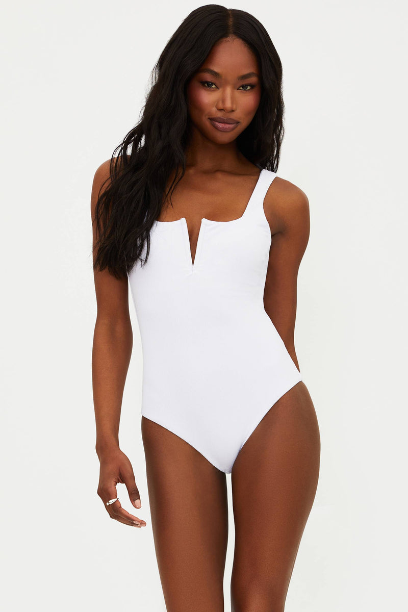 A model is wearing a white one-piece swimsuit with a ribbed texture, fixed straps, and a low back.