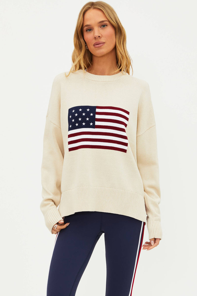 A female model wearing a white oversized crewneck sweater with a graphic of the American flag on the chest.