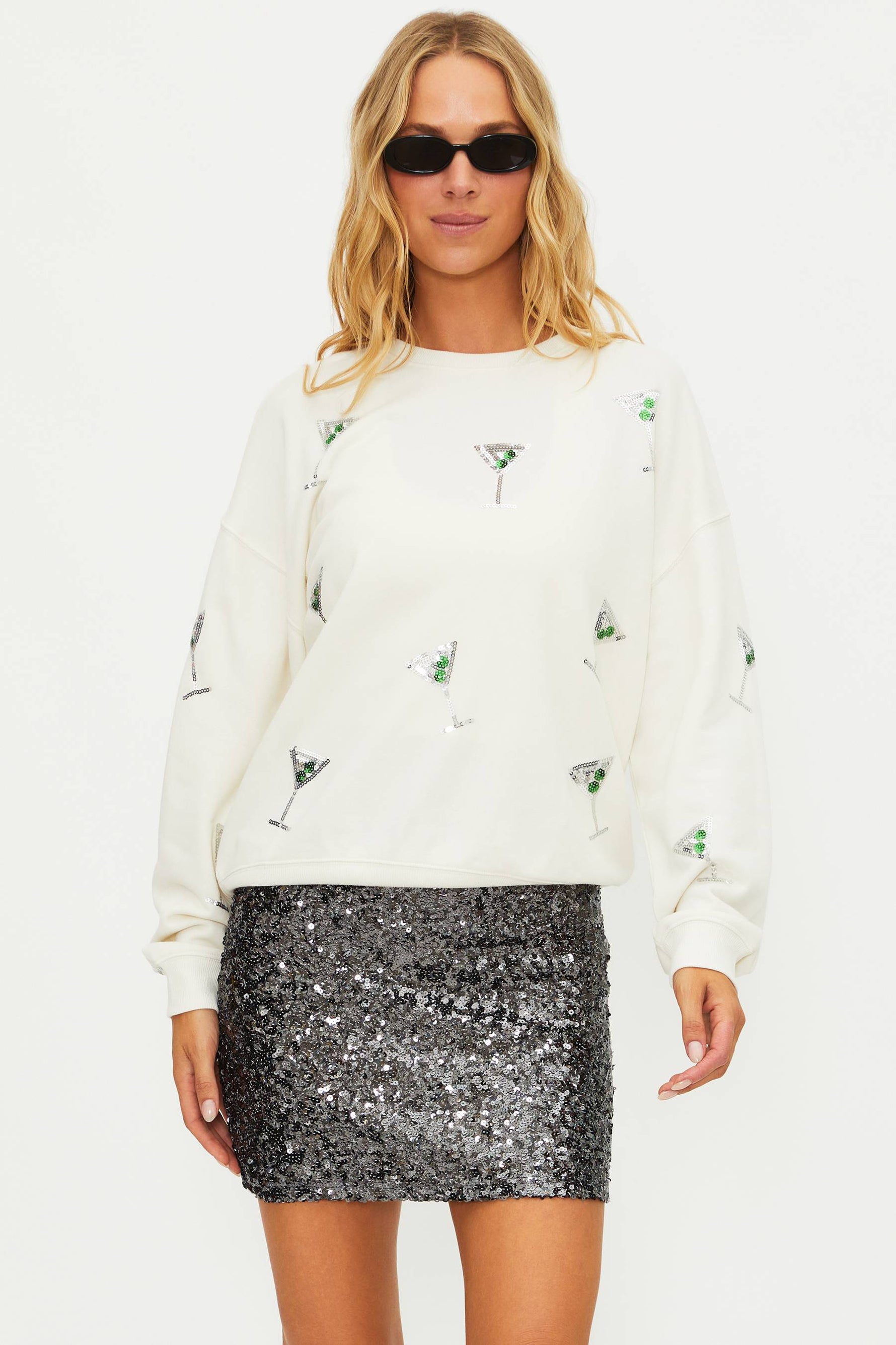 Woman wearing a white sweater with cocktail-themed designs and a silver sequin skirt on a white background