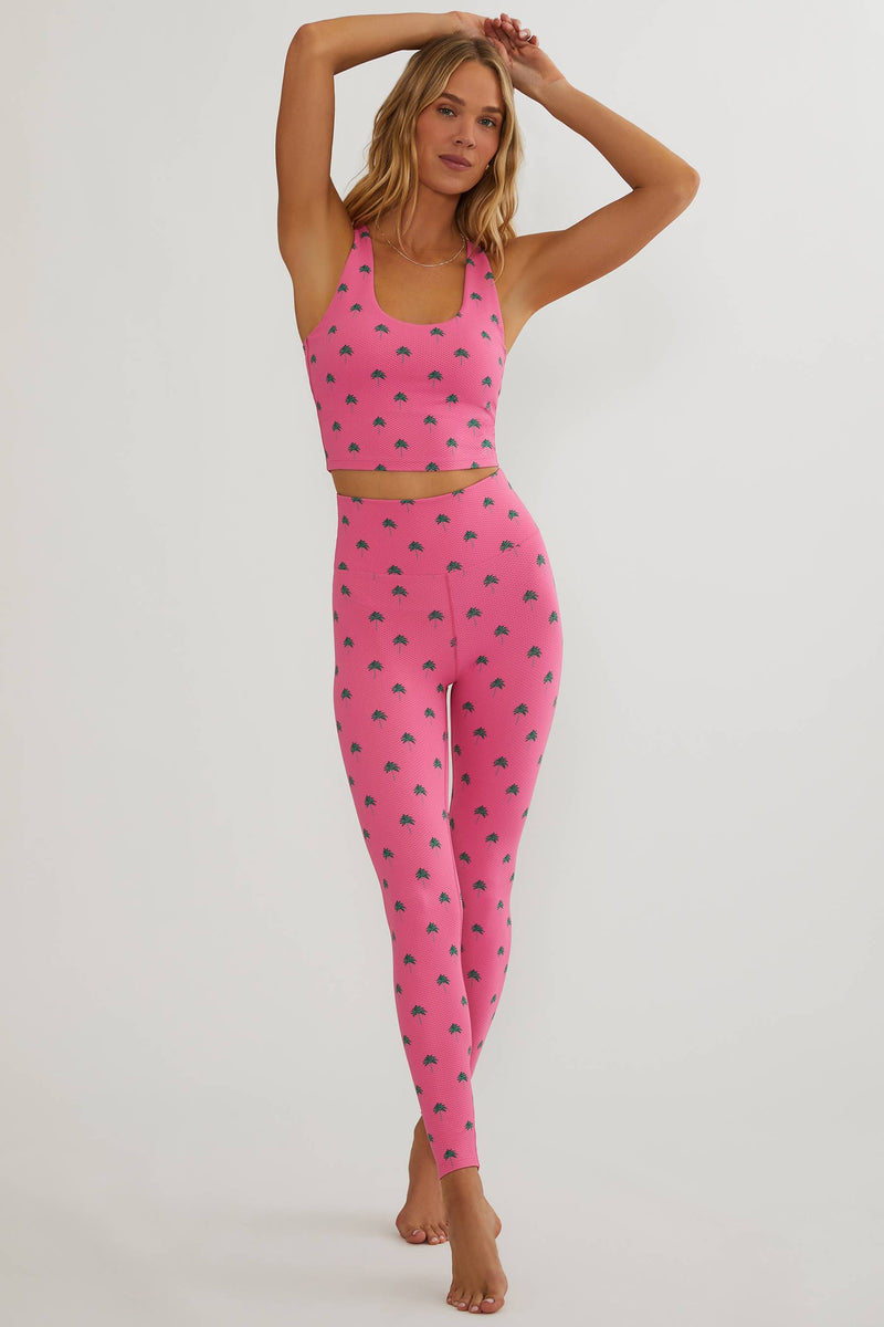 A woman modeling a matching two-piece activewear set consisting of a pink sports bra and high-waisted leggings, both featuring a repeating dark pink strawberry print, against a plain light gray studio background.