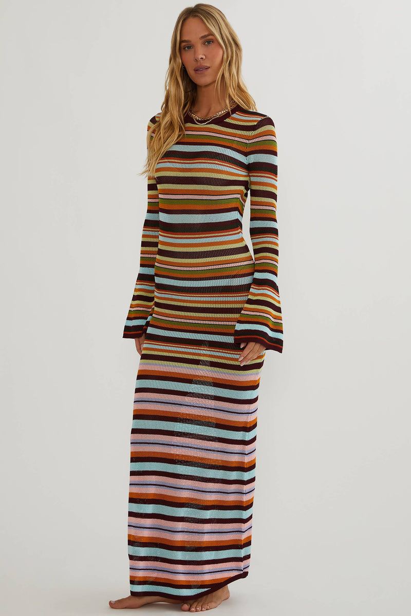 A woman modeling a long-sleeve maxi dress with horizontal multicolor stripes in brown, orange, yellow, green, blue, pink, and white, set against a plain light gray studio background.