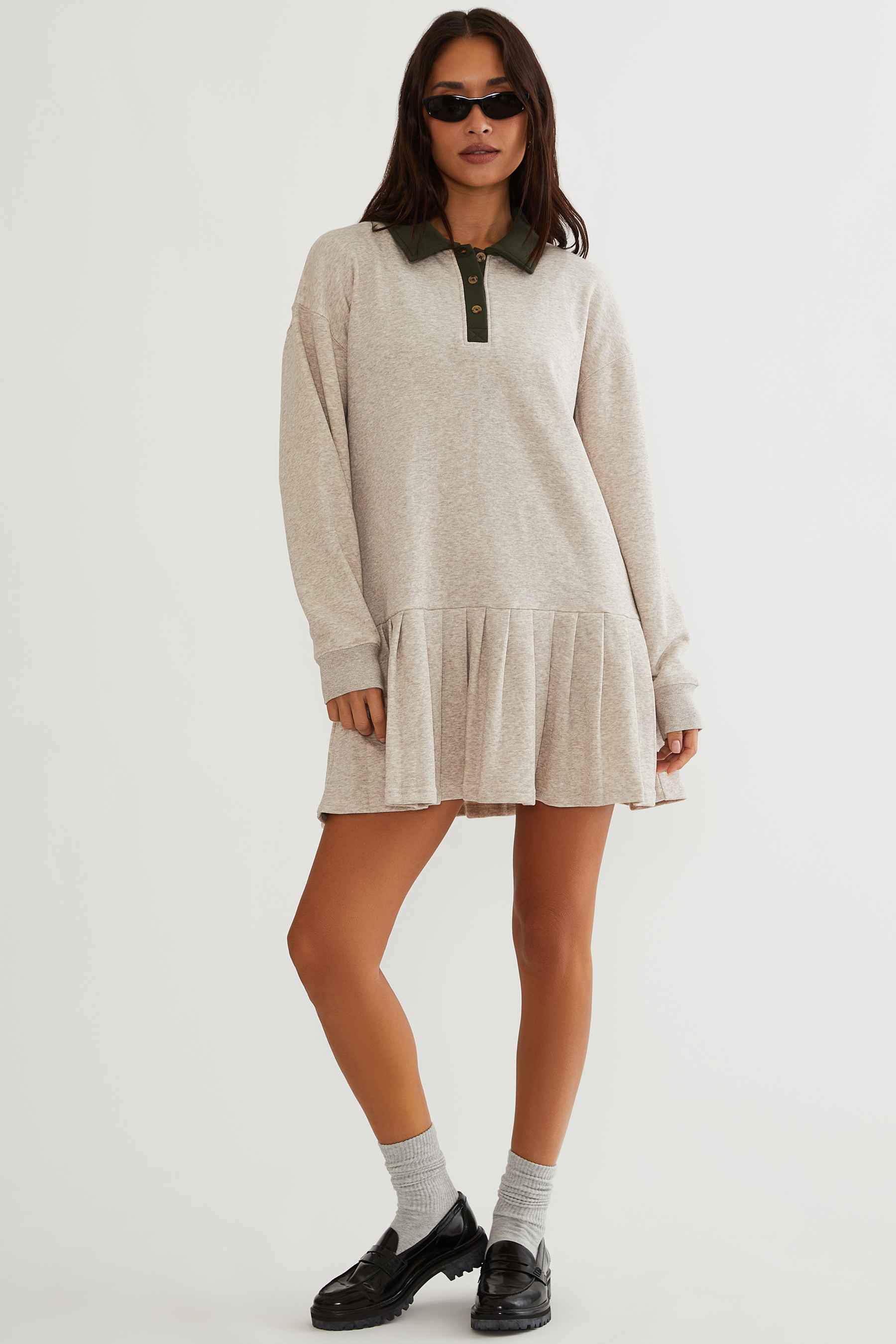 A woman modeling a light beige long-sleeve mini sweater dress with an olive green collar and placket, styled with black sunglasses, white socks, and black leather loafers against a plain white studio background.