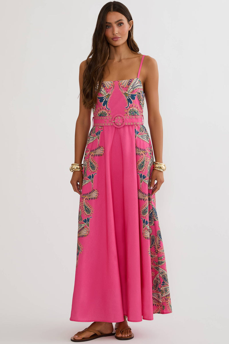 A woman modeling a bright pink maxi dress with intricate multicolor floral embroidery and a matching belt, set against a plain light gray studio background.