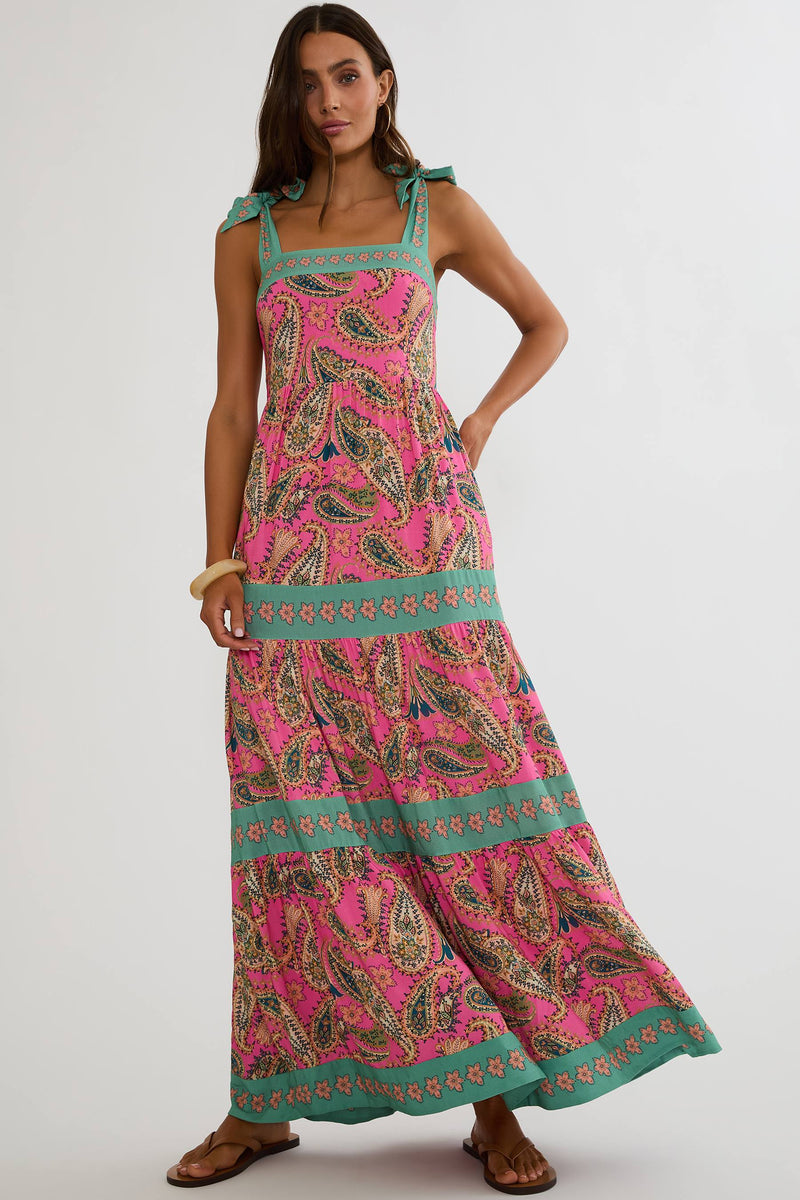 A woman modeling a sleeveless tiered maxi dress with a vibrant pink paisley pattern and turquoise green trim, shown from a three-quarters back view against a light gray studio background.