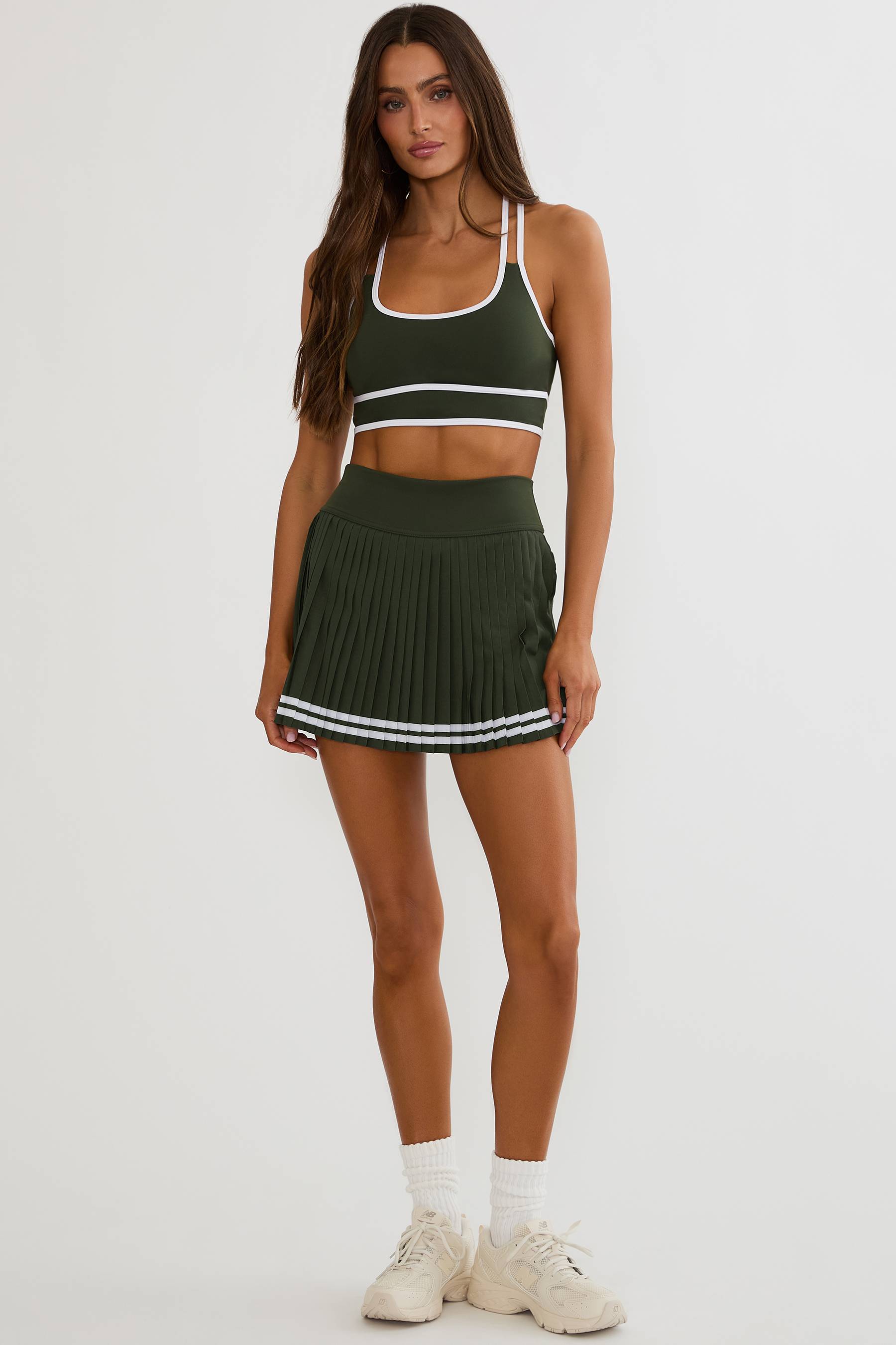A woman modeling a dark green athletic two-piece set consisting of a crop top and a pleated mini skirt, both with white trim details, paired with white socks and sneakers against a light gray studio background.