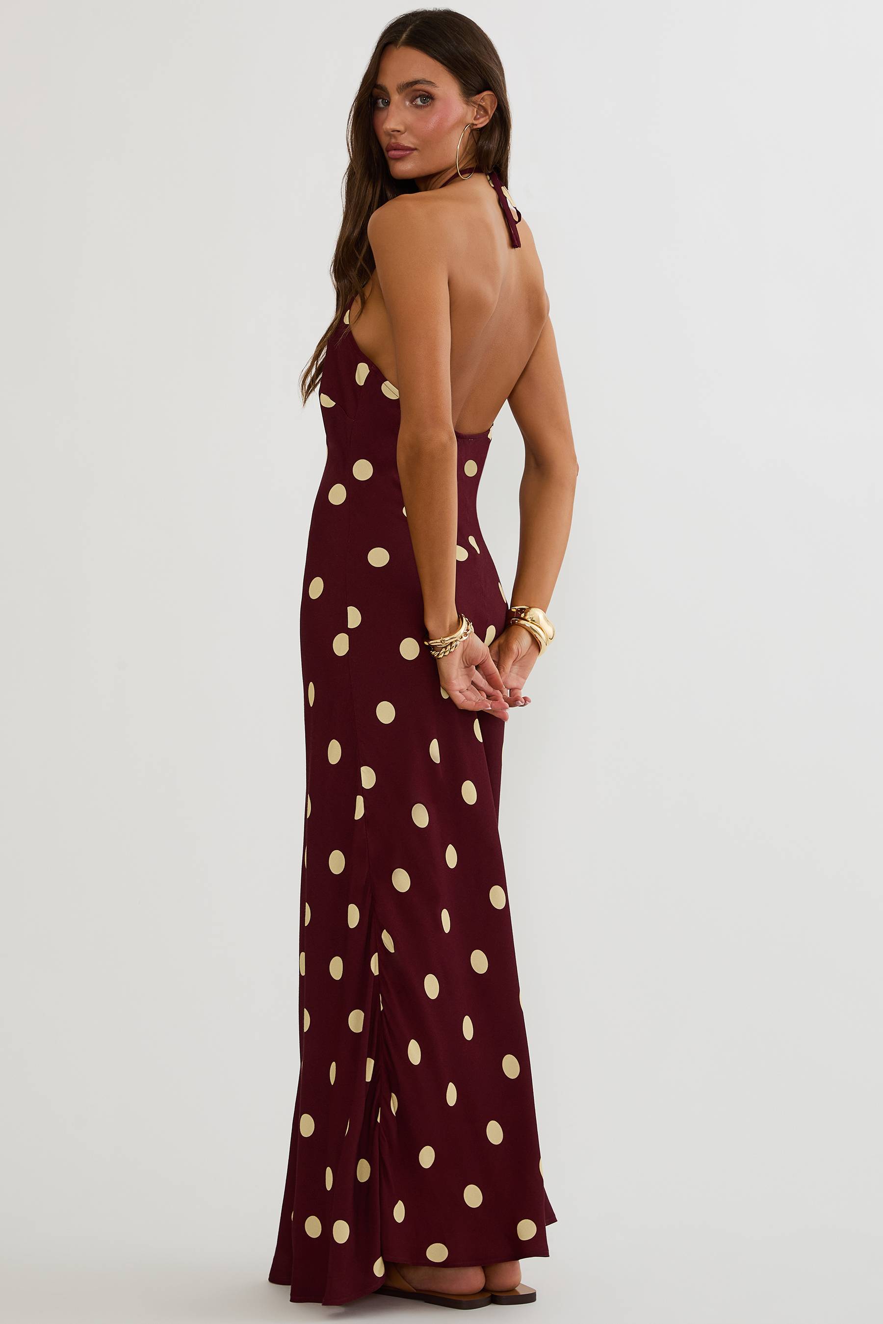 A woman modeling a sleeveless, backless maxi dress in deep burgundy with a large beige polka dot pattern, shown from a three-quarters back view against a light gray studio background.