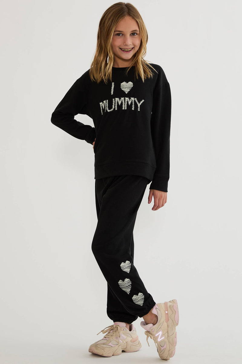 A young girl modeling a black two-piece sweatshirt and jogger set with silver glittery heart and text details, paired with beige sneakers, against a plain light gray background.