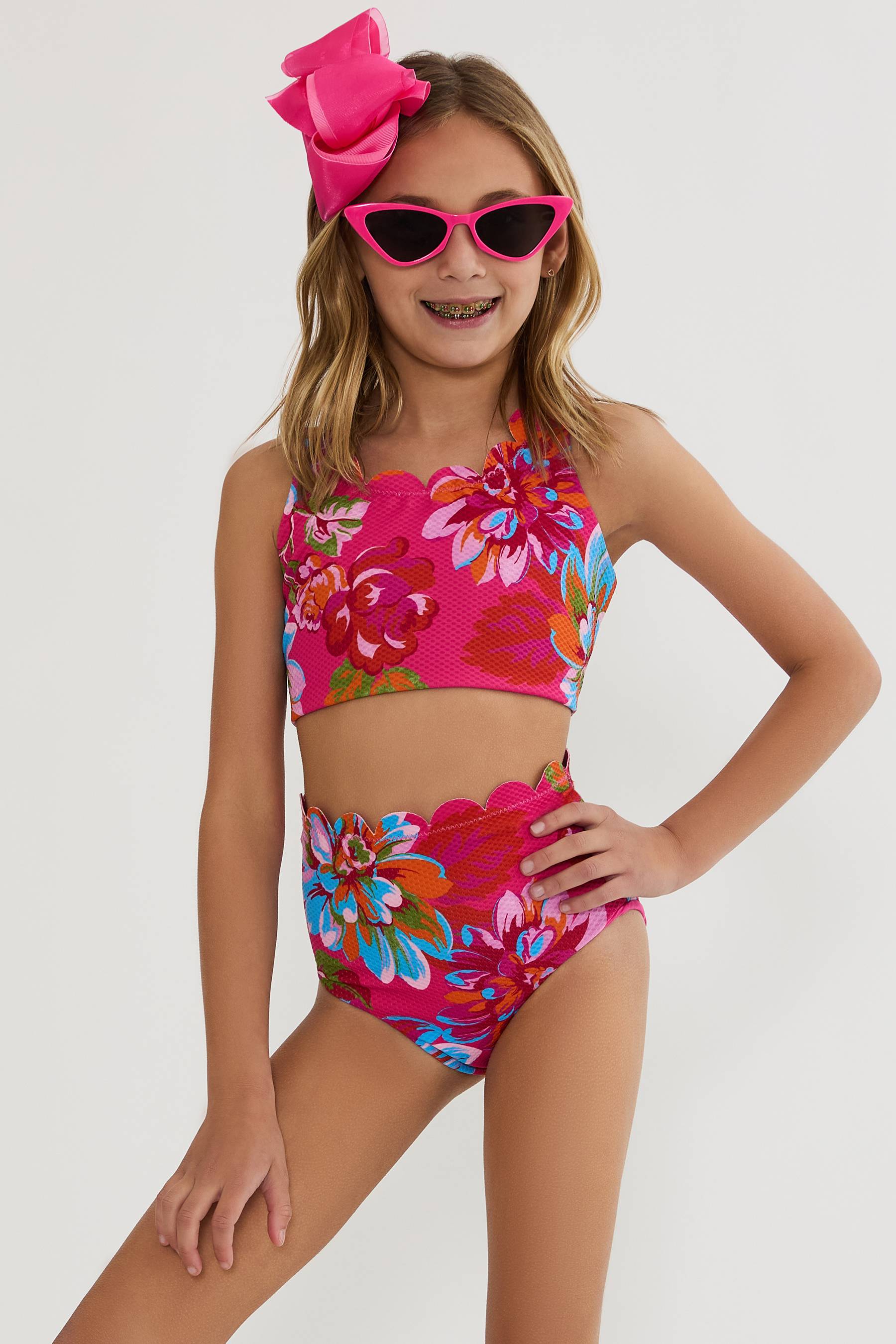 Young girl wearing a colorful floral two-piece swimsuit with pink sunglasses and headband on a white background