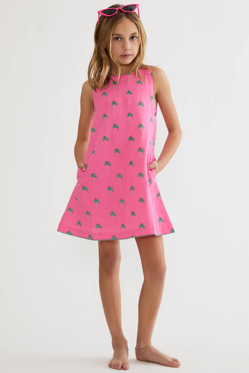A young girl stands barefoot against a plain light gray studio background, wearing a sleeveless pink dress with a small repeating green pattern and pink sunglasses on her head.