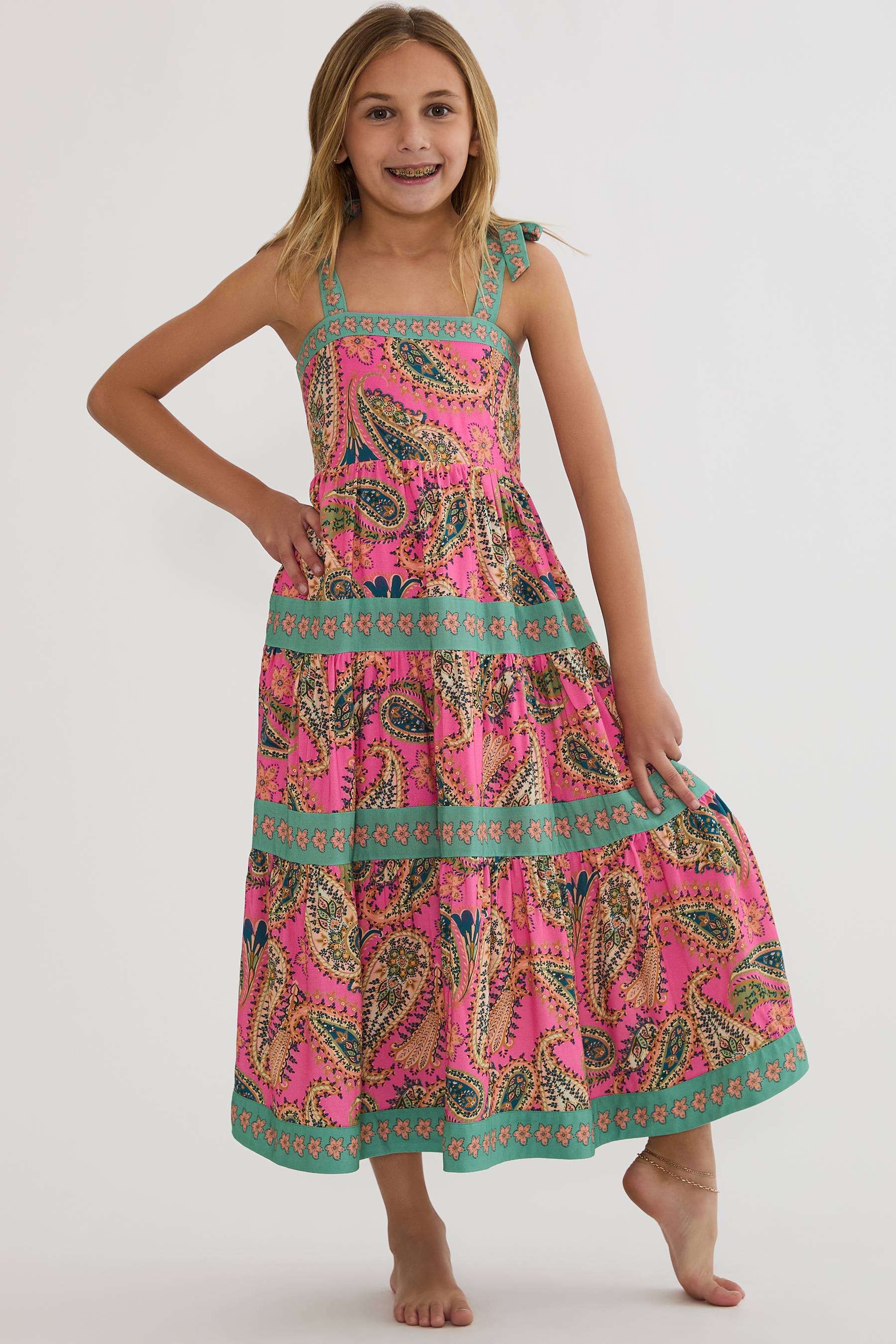 A young girl modeling a sleeveless tiered maxi dress with a vibrant pink paisley pattern and teal borders, set against a plain light gray studio background.