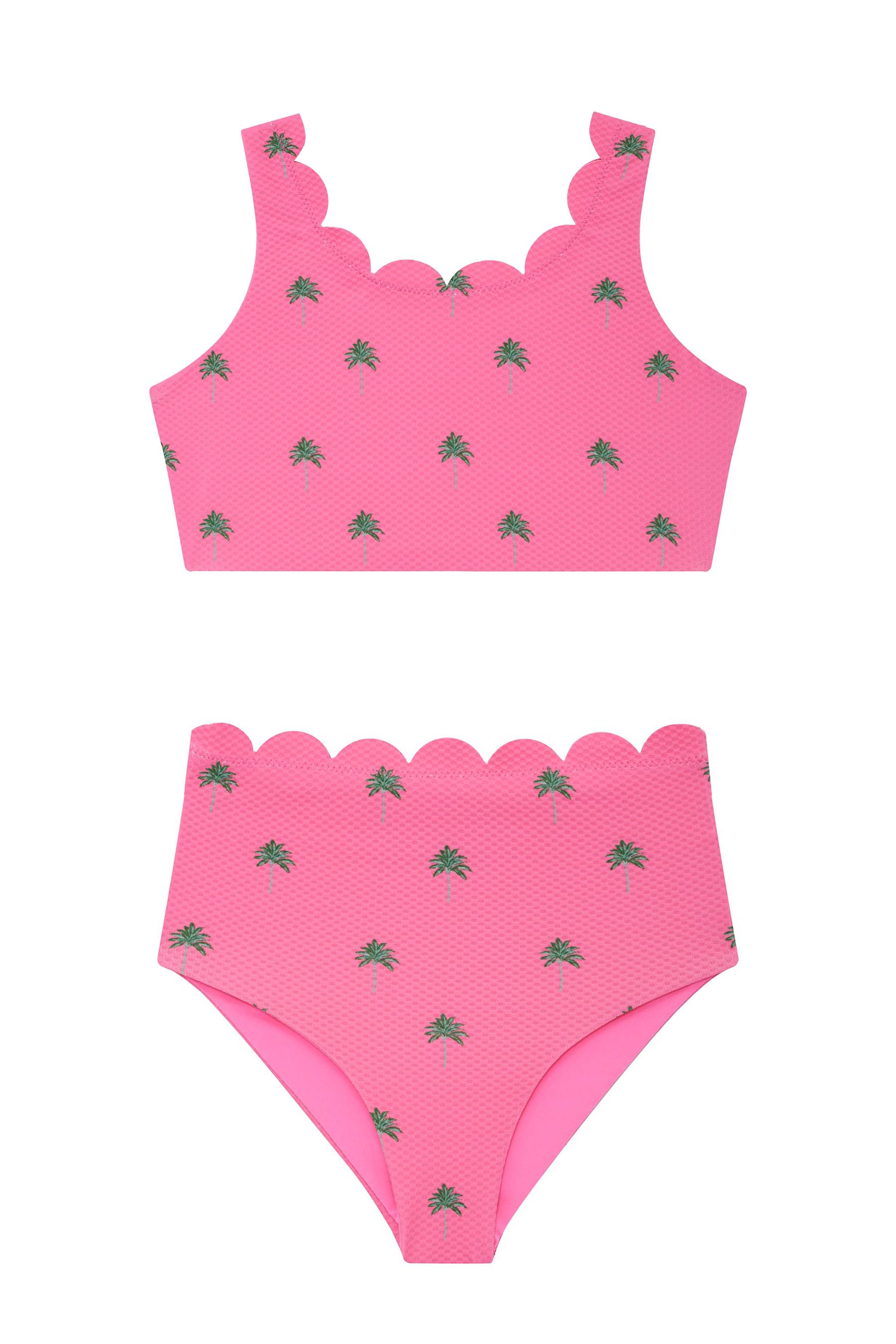 A young girl modeling a bright pink two-piece swimsuit with a small green leaf pattern, matching pink sunglasses, and a large pink bow headband against a light gray studio background.