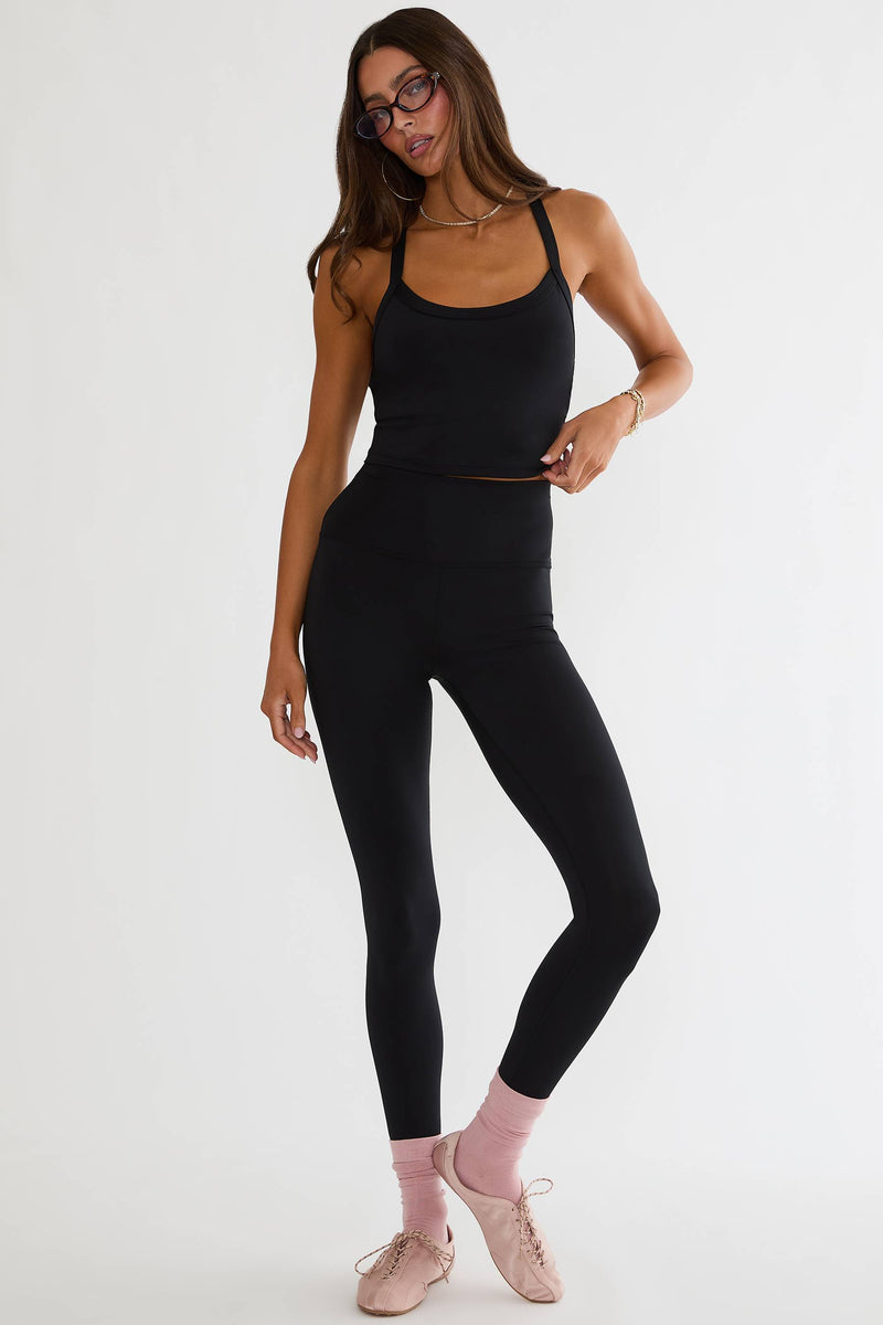 A woman modeling a black activewear set consisting of a scoop-neck tank top and high-waisted leggings, paired with light pink socks and ballet flats, against a plain white studio background.
