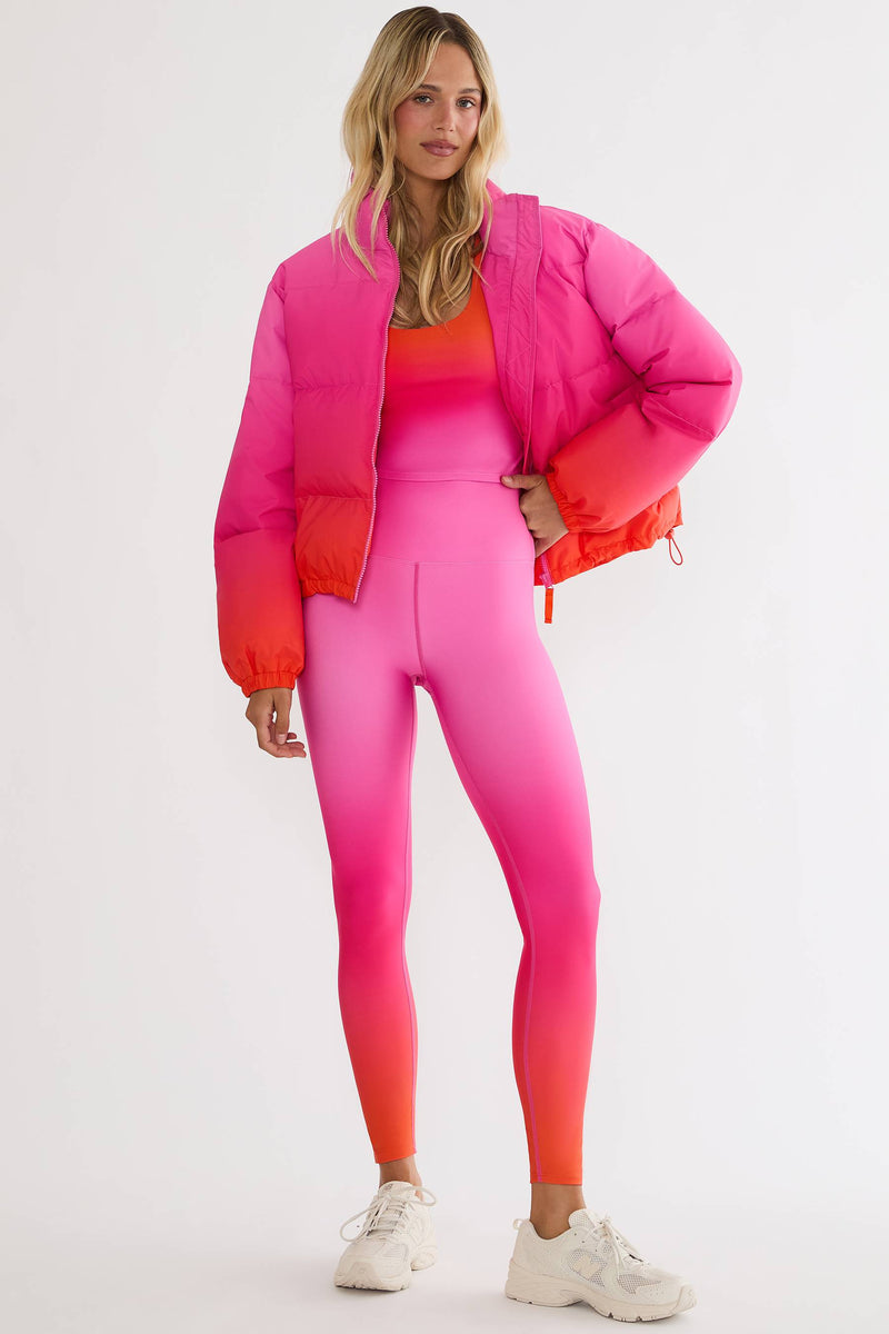 A woman modeling a vibrant pink and orange gradient puffer jacket over a matching pink and orange gradient activewear set, paired with white sneakers, against a plain white studio background.