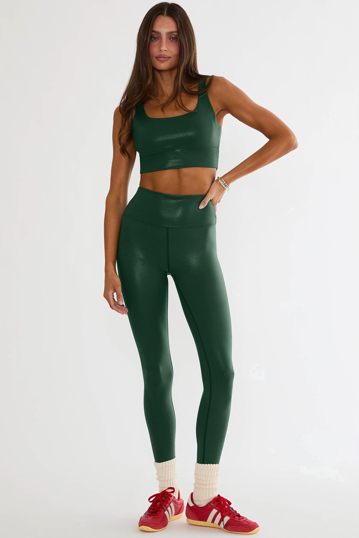 A woman modeling a matching dark green faux-leather activewear set consisting of a cropped tank top and high-waisted leggings, paired with red sneakers and cream socks, against a plain white studio background.