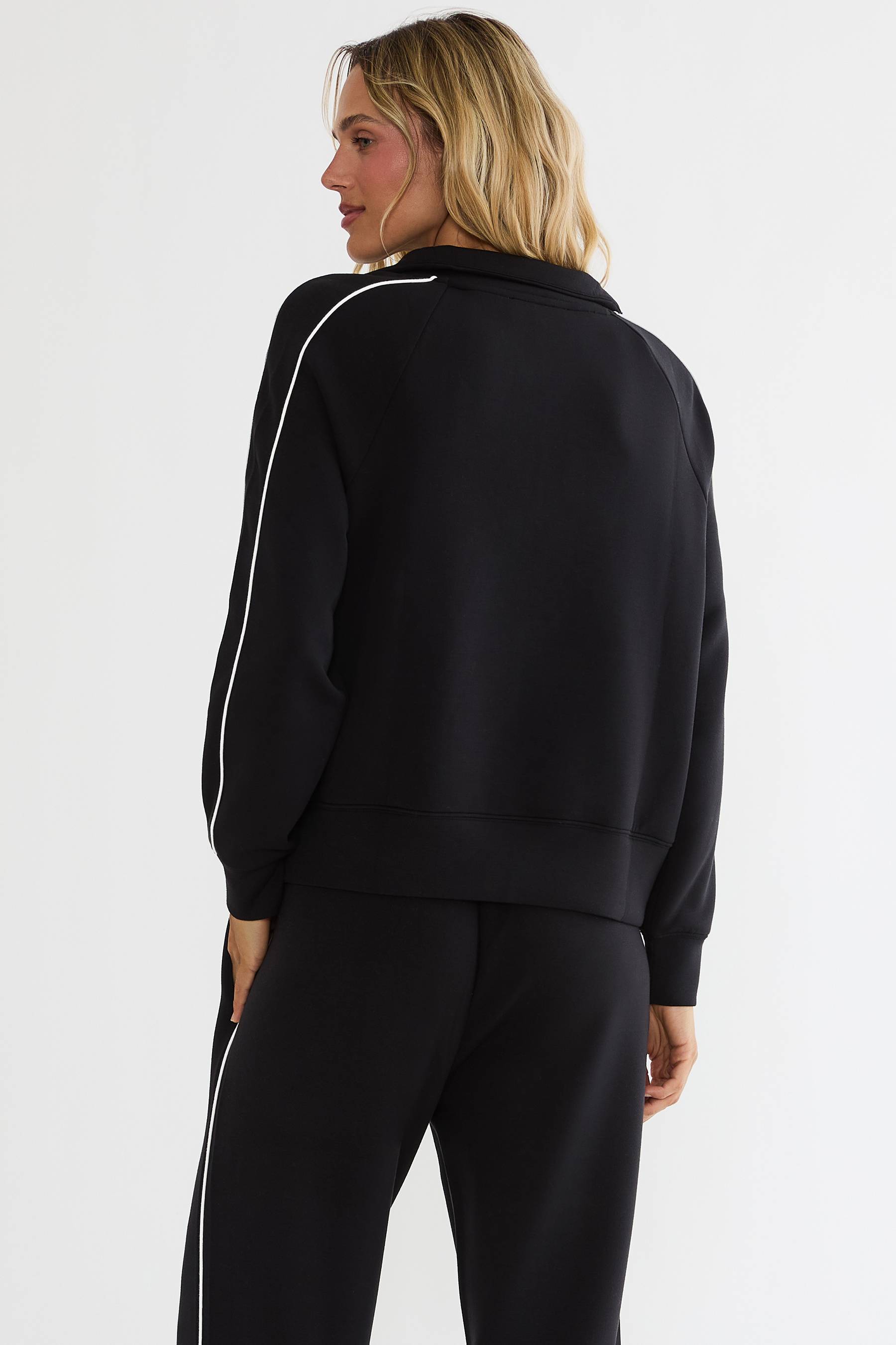 A woman shown from the back wearing a black cropped zip-up sweatshirt with white piping along the sleeve and matching black sweatpants with white piping, against a plain white studio background.