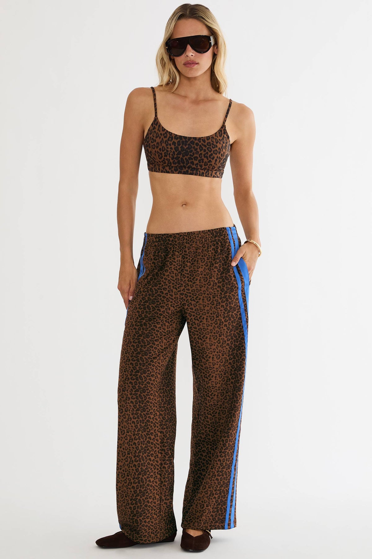 A model wearing a matching two-piece leopard print set with wide-leg pants featuring blue side stripes, a leopard print crop top, and dark sunglasses, photographed against a plain white studio background.