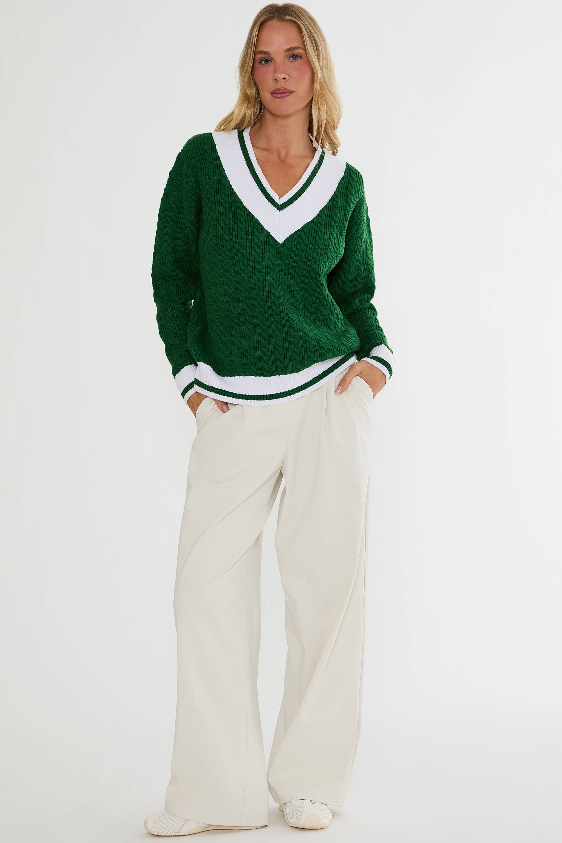 Woman wearing a green sweater with a white V-neck and white pants on a light gray background