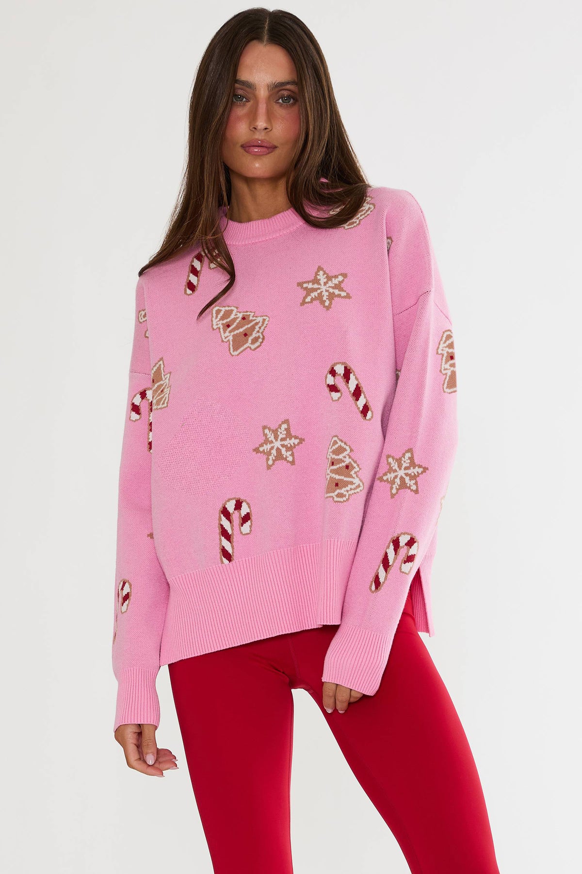 A woman modeling a pink knit sweater with festive Christmas motifs including gingerbread cookies, candy canes, and stars, paired with red leggings against a plain light gray studio background.