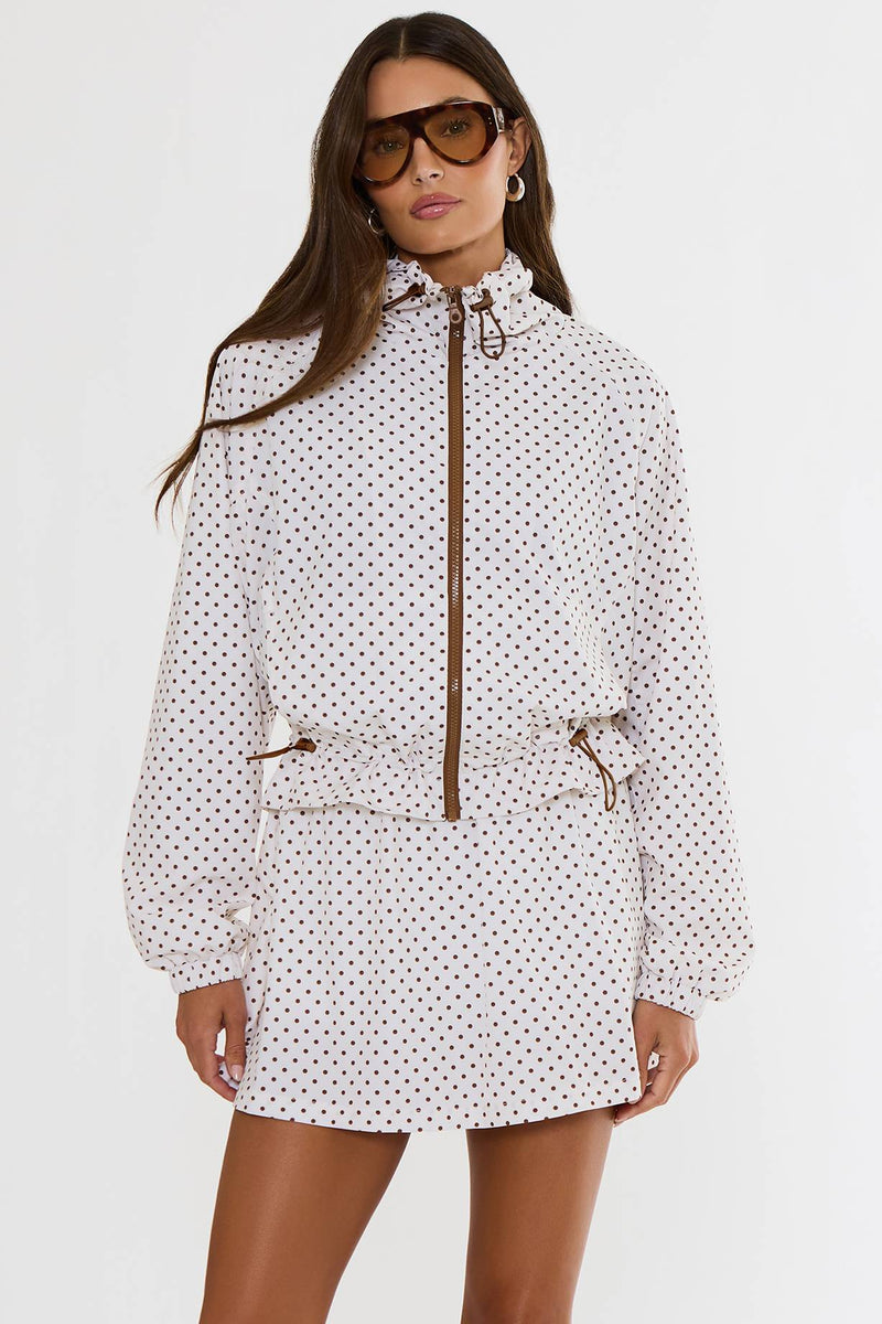 A model wearing a white long-sleeve hooded jacket and matching mini skirt set, both decorated with a small red polka dot pattern, featuring brown zipper and drawstring details. She is also wearing brown sunglasses against a plain white studio background.
