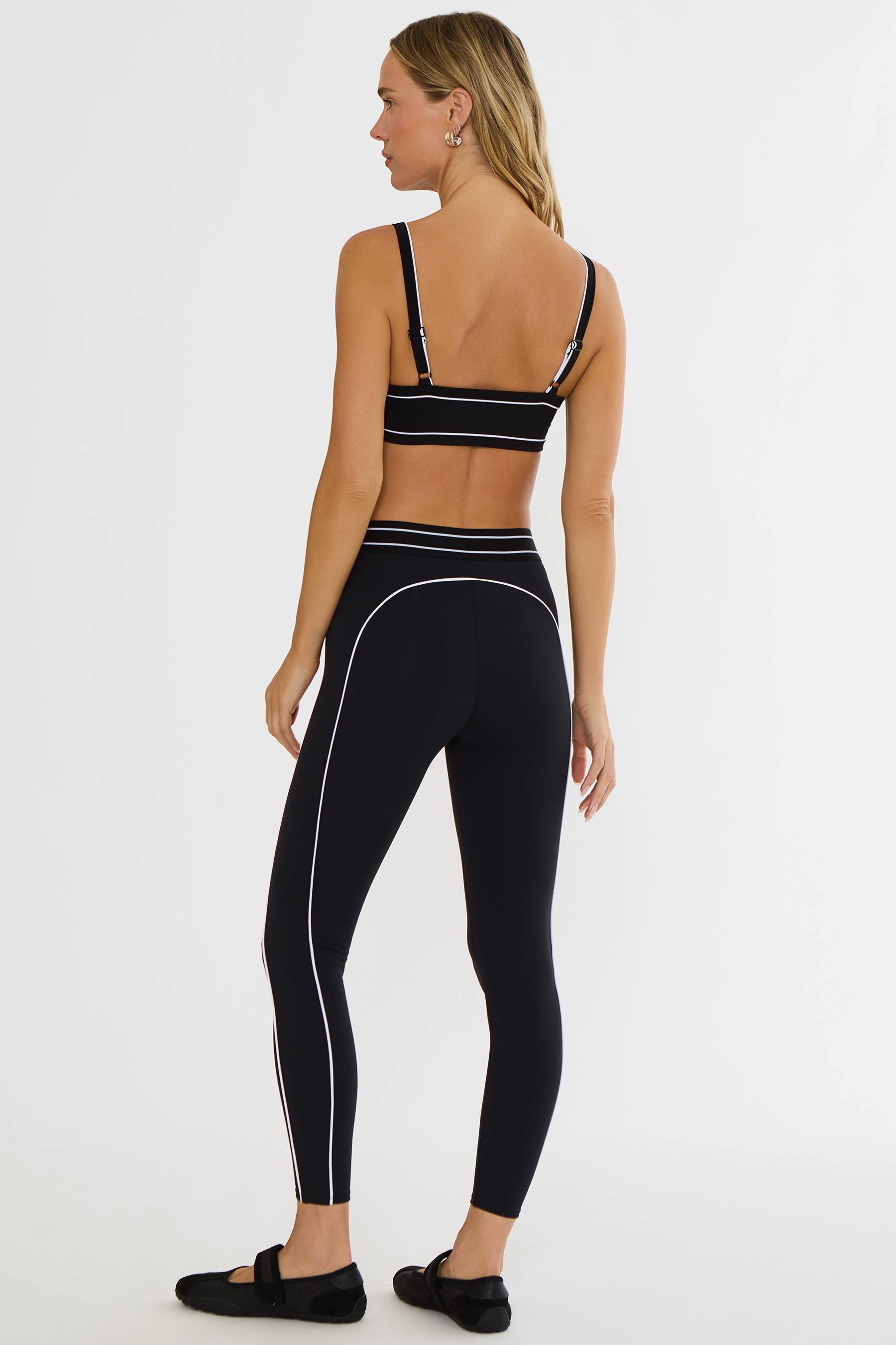 A woman shown from the back wearing a matching black activewear set with white contrast piping, consisting of a sports bra and high-waisted leggings, against a plain white studio background.