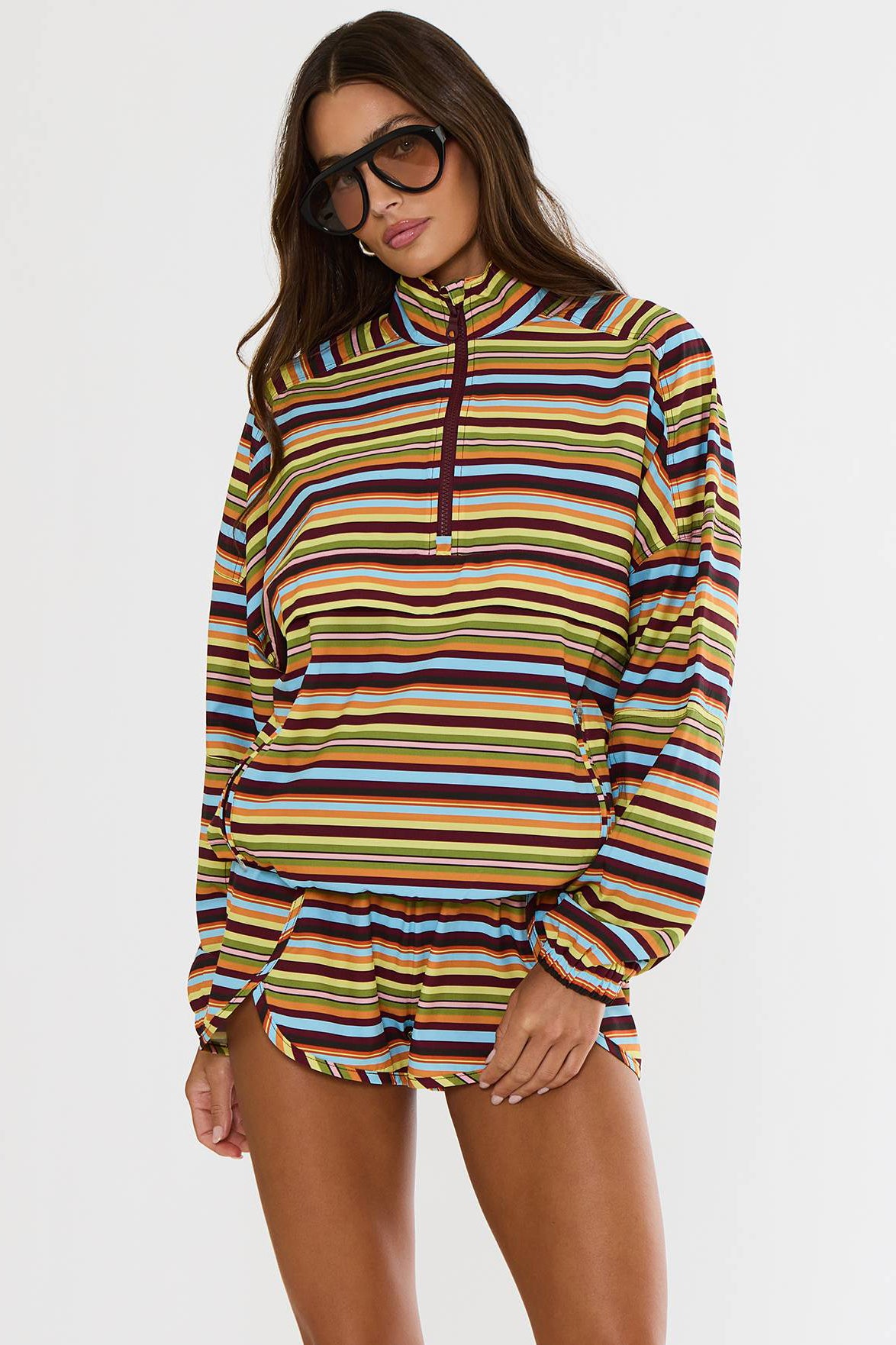 A woman modeling a long-sleeve quarter-zip pullover top with horizontal multicolor stripes in yellow, orange, brown, blue, and white, set against a plain light gray studio background.