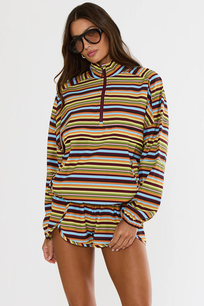A woman modeling a long-sleeve quarter-zip pullover top with horizontal multicolor stripes in yellow, orange, brown, blue, and white, set against a plain light gray studio background.
