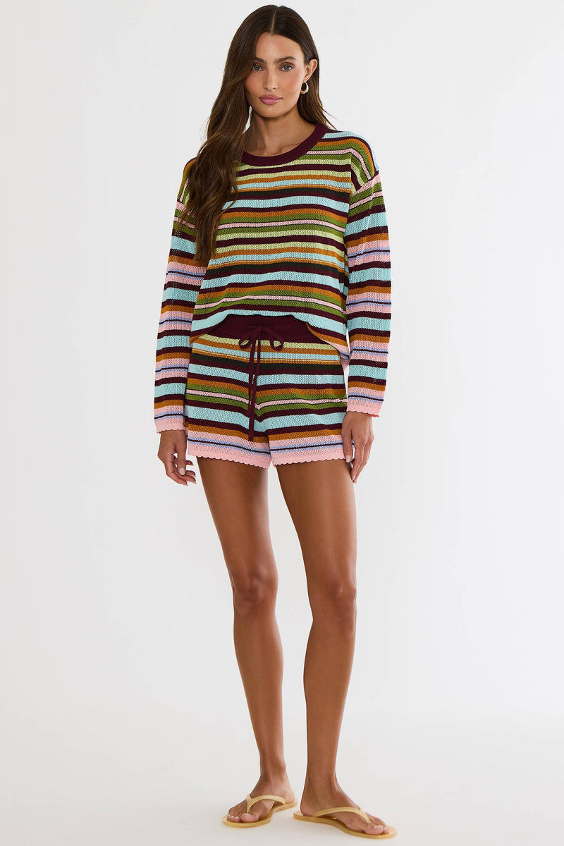 A woman modeling a long-sleeve knit top and matching shorts set with horizontal stripes in brown, green, orange, pink, blue, and white, featuring a dark burgundy drawstring waist, on a light gray studio background.