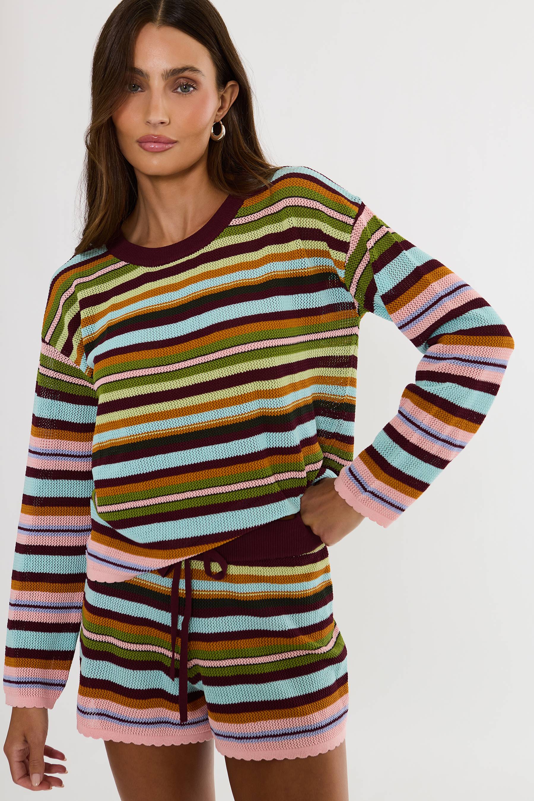 A woman modeling a matching striped knit long-sleeve top and shorts set. The set features horizontal stripes in pink, light blue, olive green, mustard yellow, brown, and dark burgundy, with a solid dark burgundy crew neck and drawstring waist.