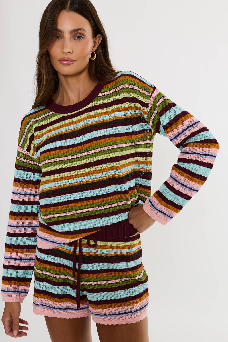 A woman modeling a matching striped knit long-sleeve top and shorts set. The set features horizontal stripes in pink, light blue, olive green, mustard yellow, brown, and dark burgundy, with a solid dark burgundy crew neck and drawstring waist.