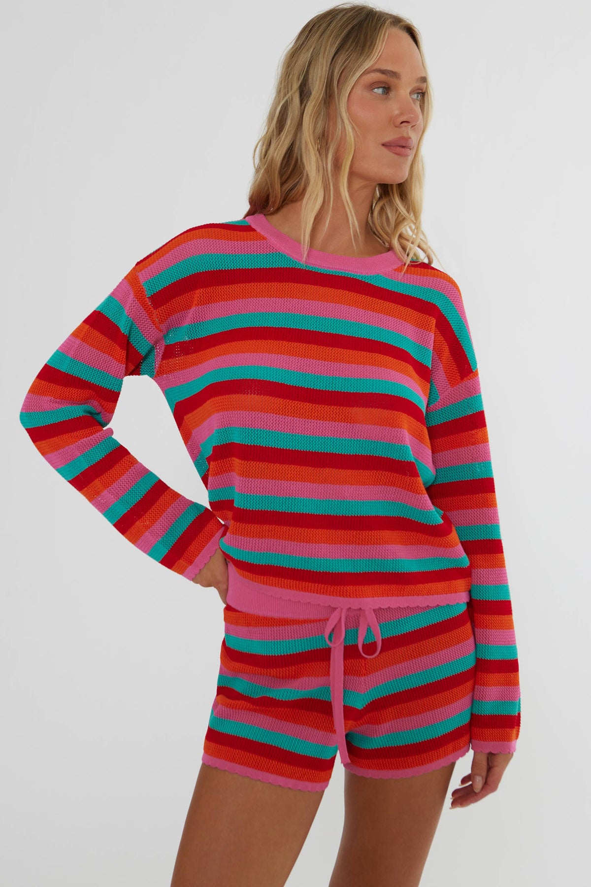Woman wearing a crochet long sleeve and short set with red, pink, orange, and teal stripes. 