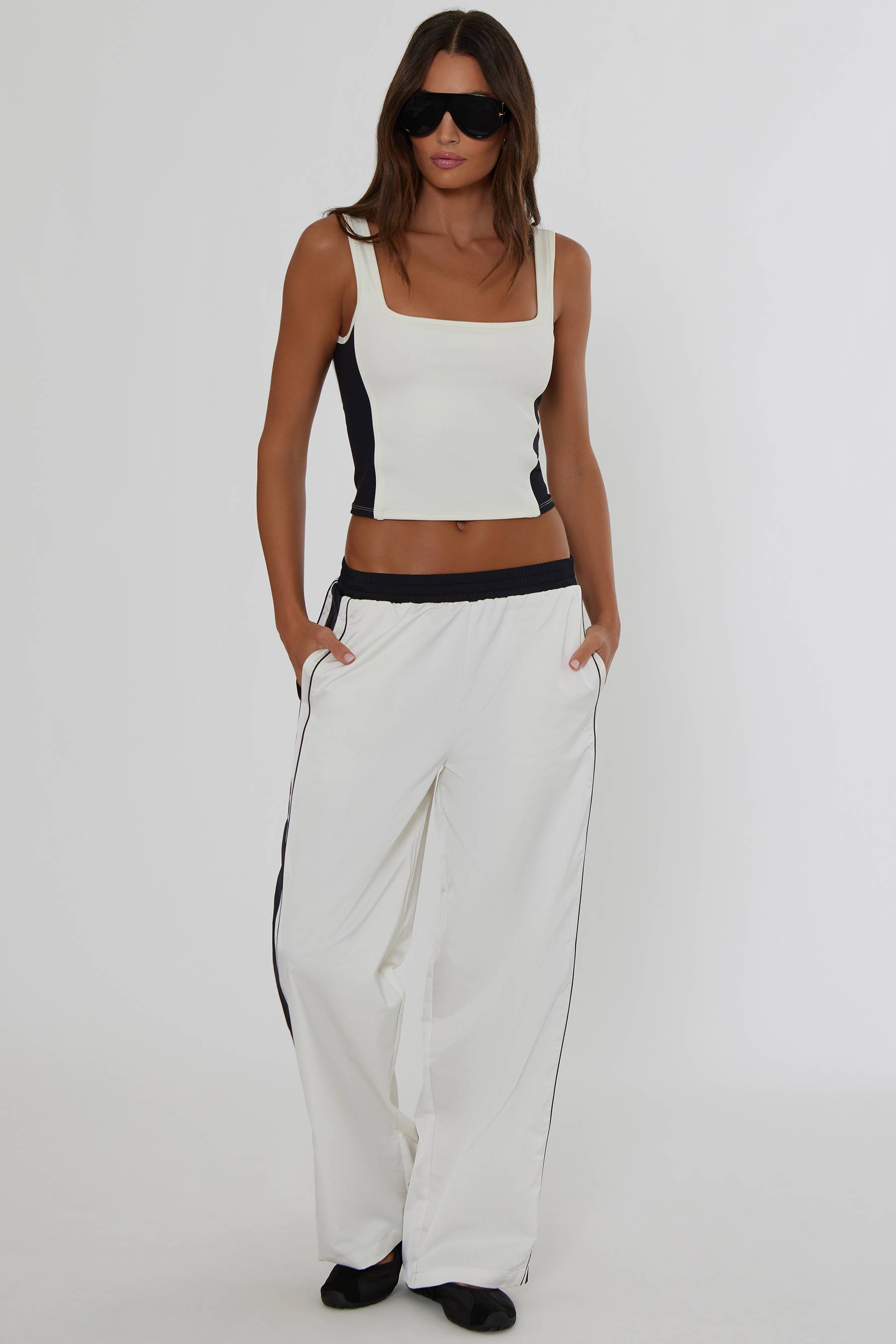 A woman modeling a white two-piece outfit consisting of a cropped tank top with black side panels and matching wide-leg sweatpants with black side stripes, set against a plain light gray studio background.