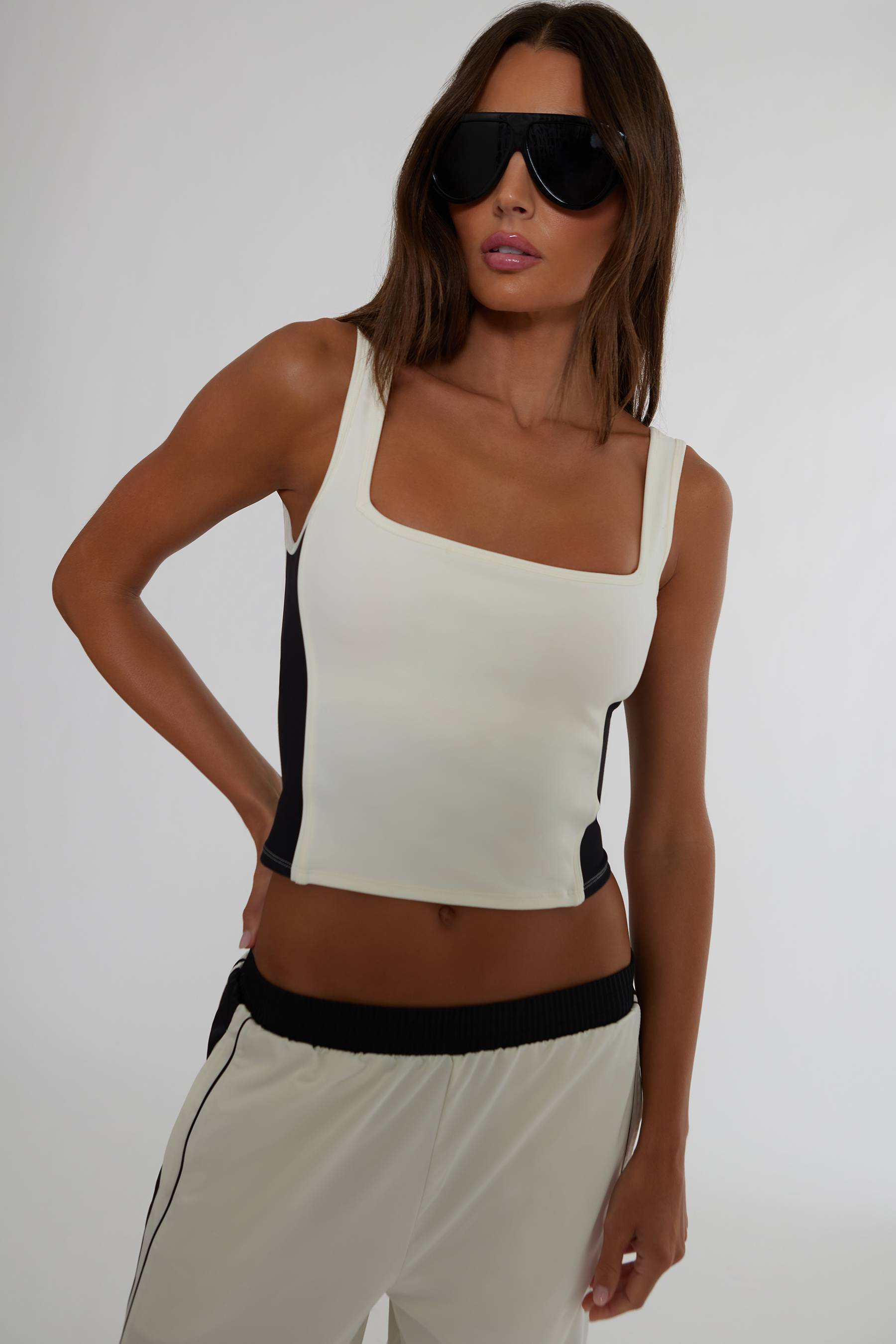 A woman modeling a fitted sleeveless crop top in off-white with black side panels, paired with matching off-white pants with a black waistband, against a plain light gray studio background.