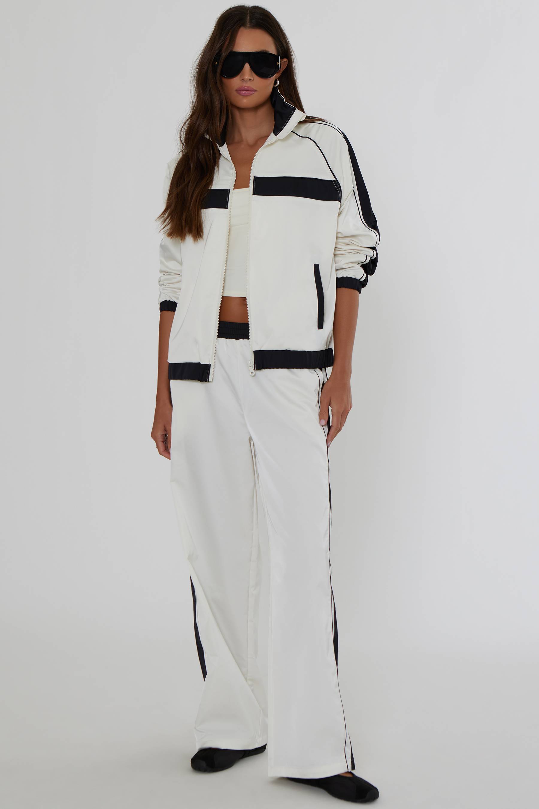 A woman modeling a matching white tracksuit with bold black stripes, featuring a zip-up jacket, cropped top, and wide-leg pants, styled with black sunglasses and shoes against a plain light gray studio background.