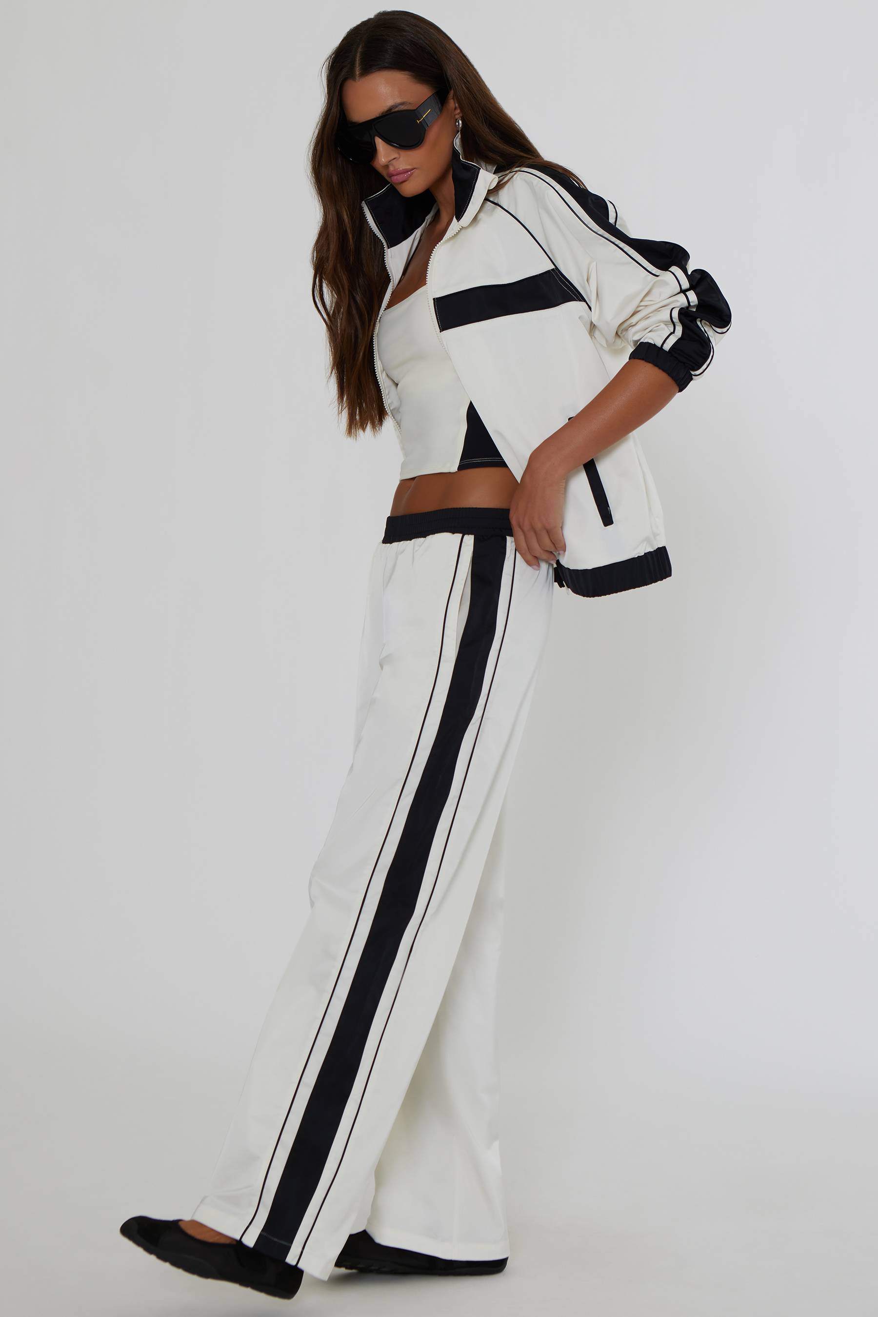 A woman modeling a matching white and black athletic tracksuit, featuring a cropped zip-up jacket and wide-leg pants with black side stripes, set against a plain light gray studio background.