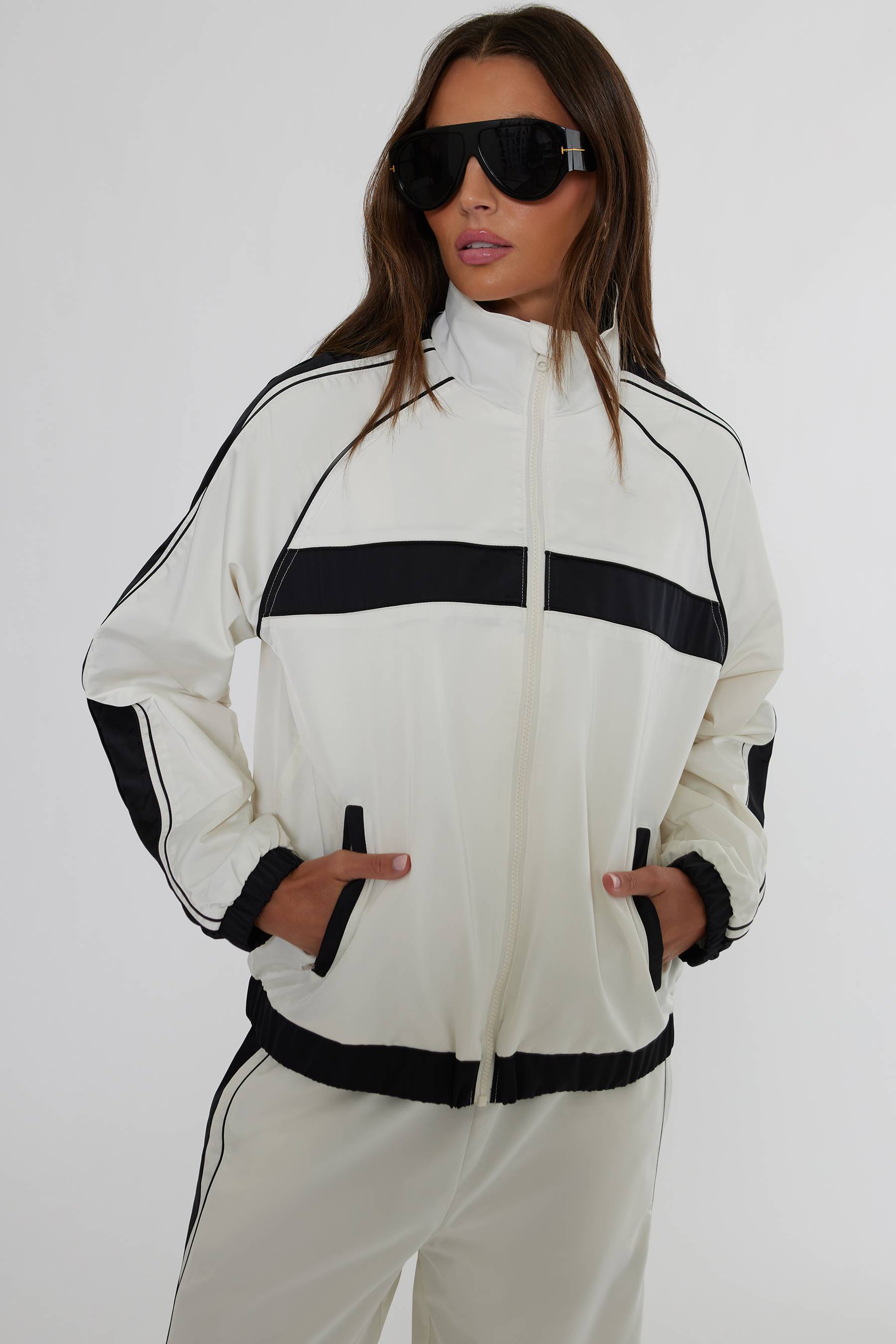 A woman modeling a white track jacket with bold black horizontal stripes and black trim, paired with matching white track pants against a plain light gray studio background.