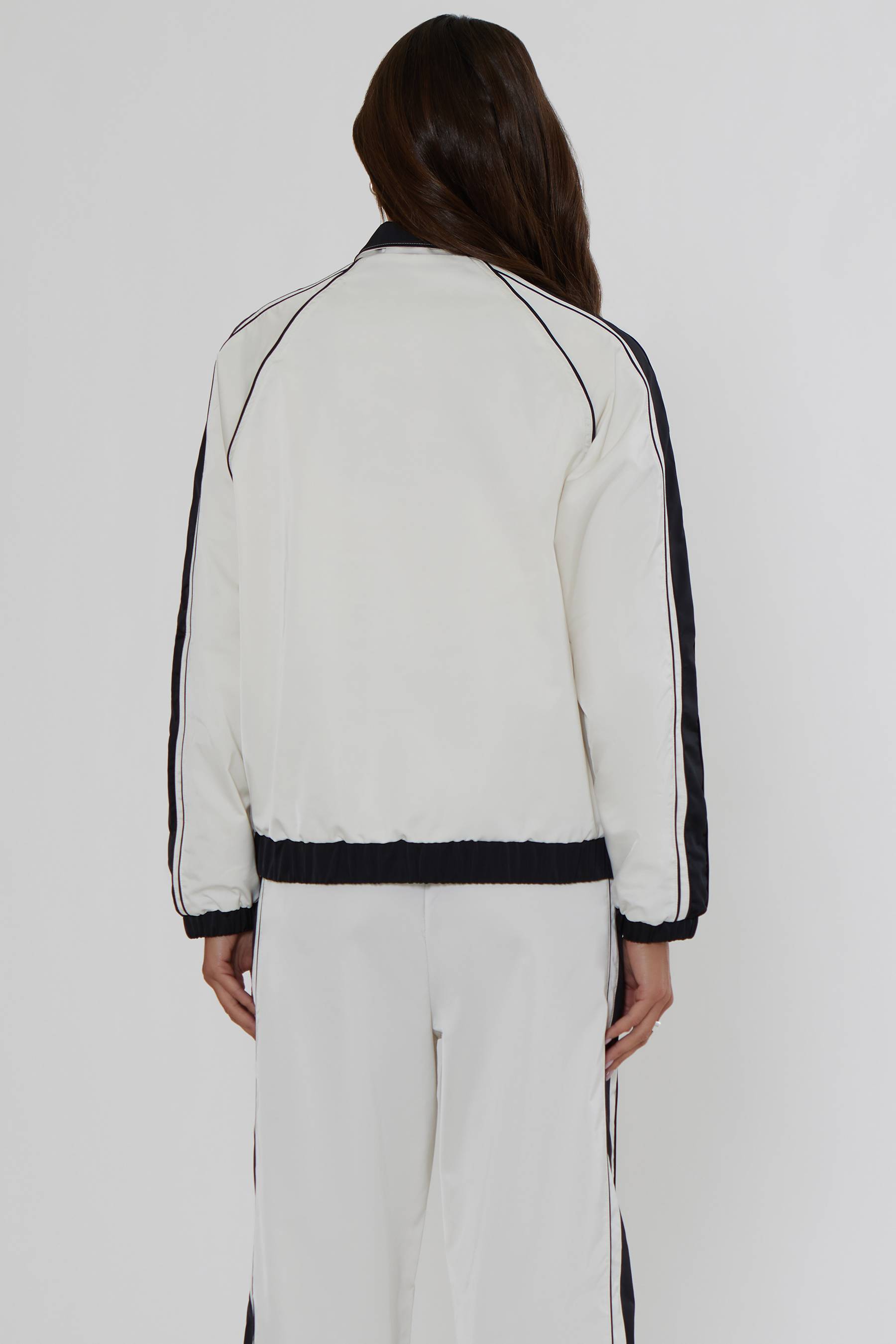 A model viewed from the back wearing a white cropped track jacket with black contrast piping and black side stripes, paired with matching white track pants against a light gray studio background.