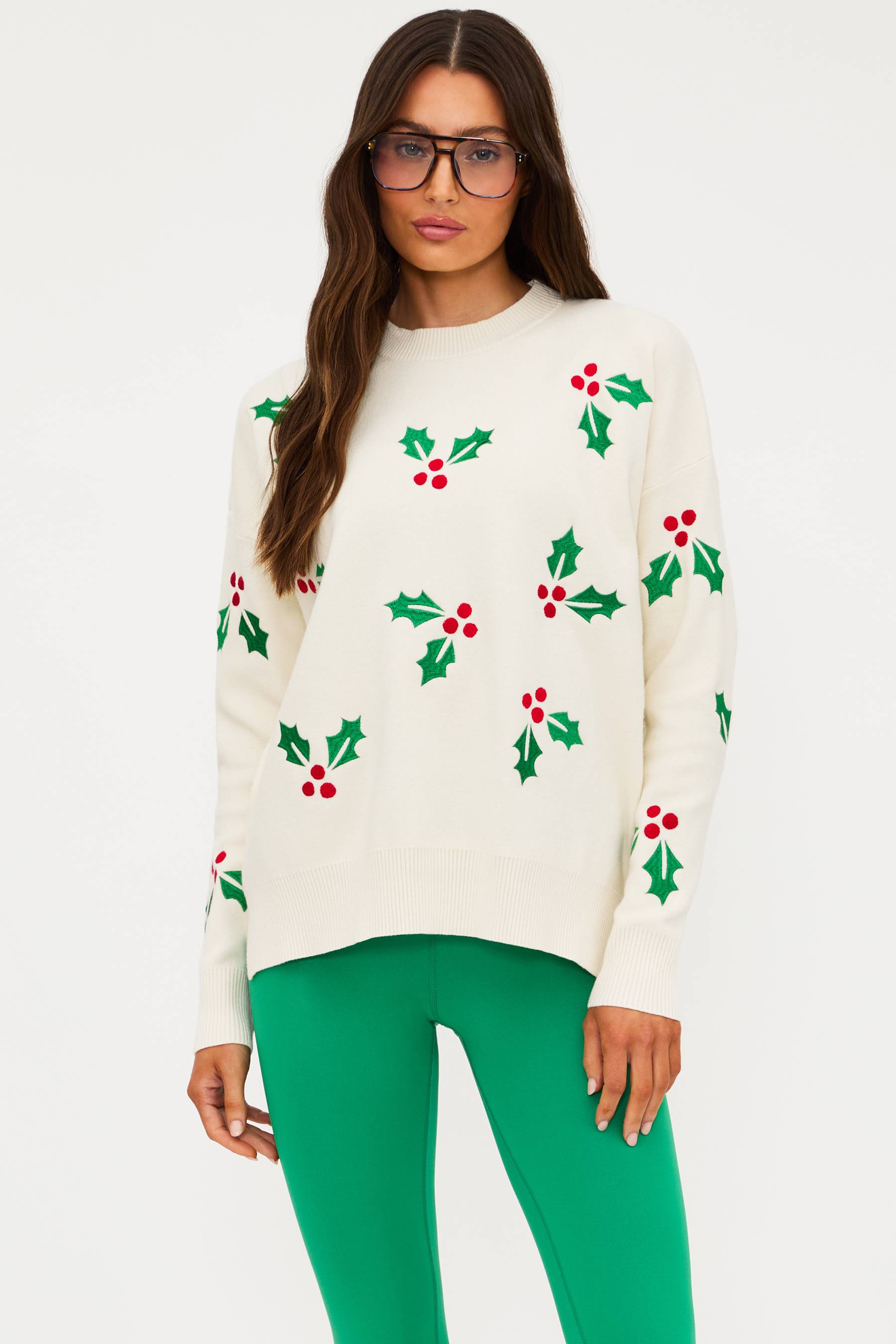 Woman wearing a white sweater with holly pattern and green pants on a white background