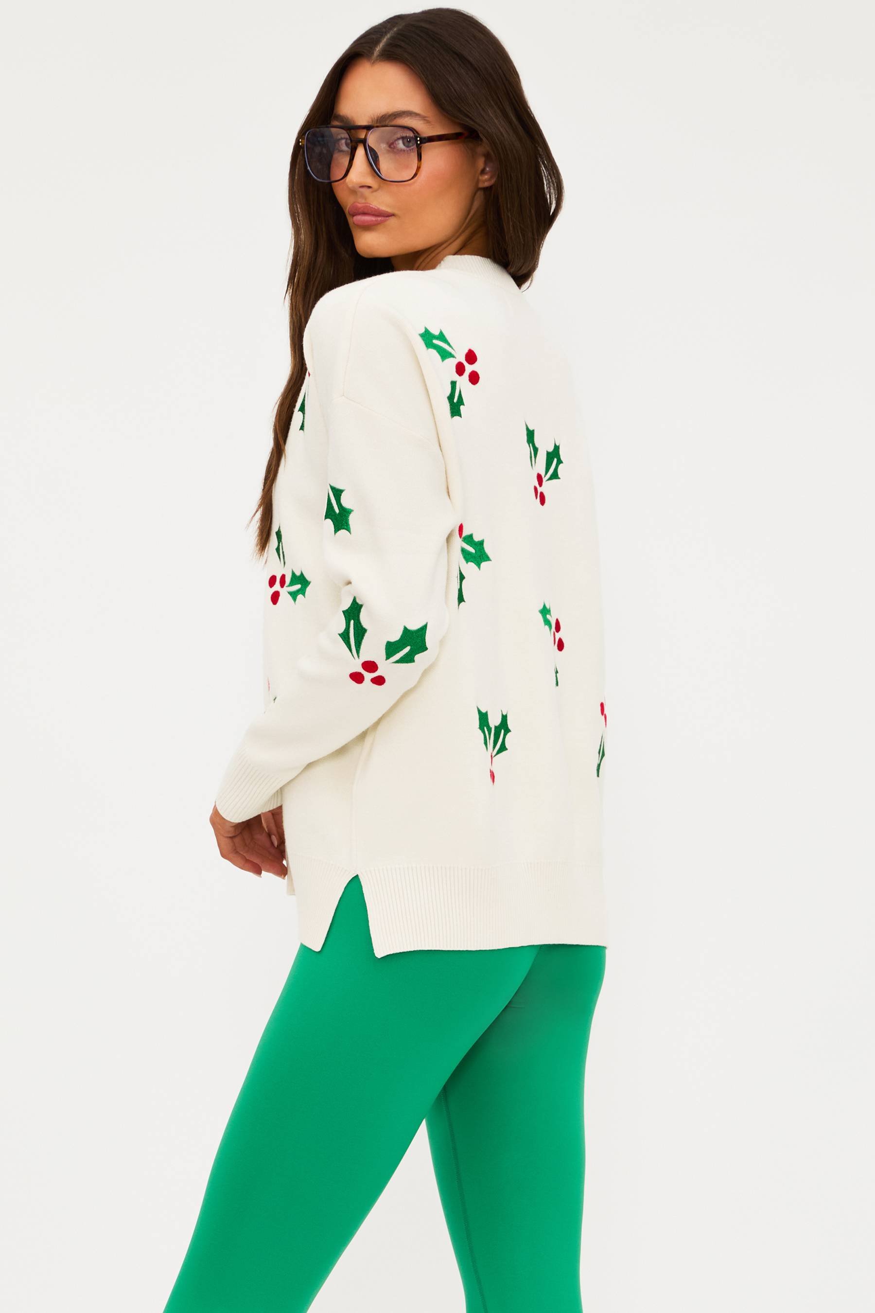 Woman wearing a white sweater with holly pattern and green pants on a white background