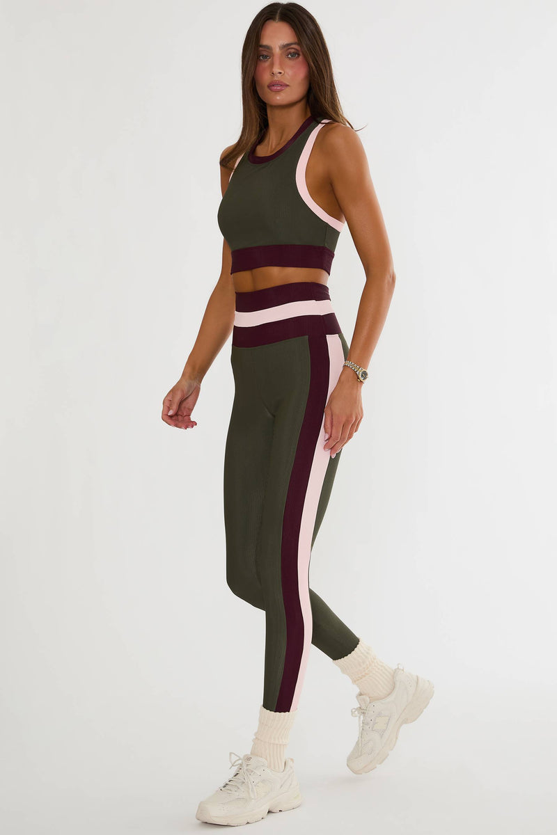 A woman modeling a matching two-piece activewear set featuring an olive green crop top and leggings with contrasting burgundy and white stripes, paired with white sneakers against a plain white studio background.